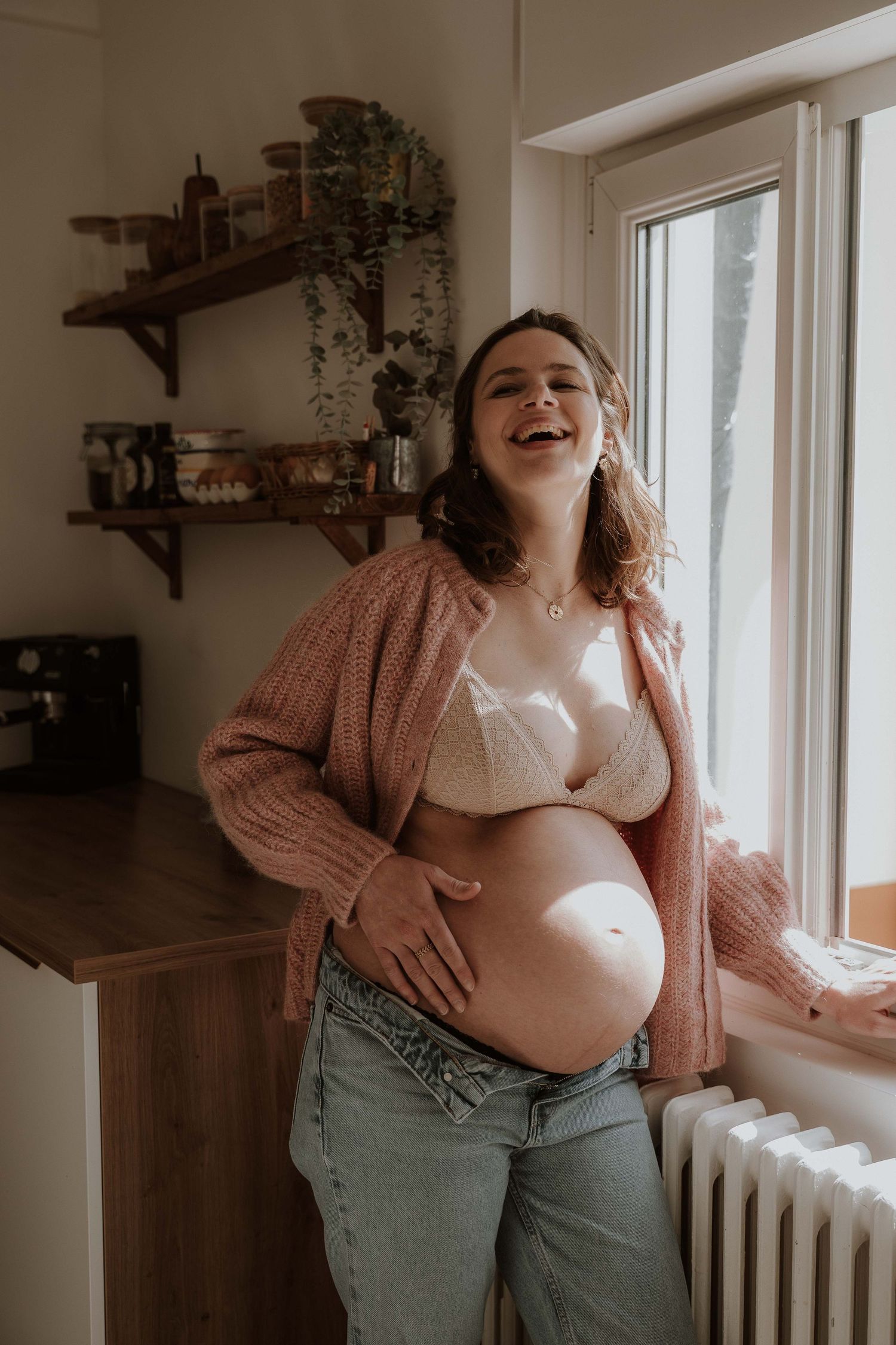 Smiling pregnant woman in pink cardigan and jeans holding baby bump near sunny window in cozy kitchen.