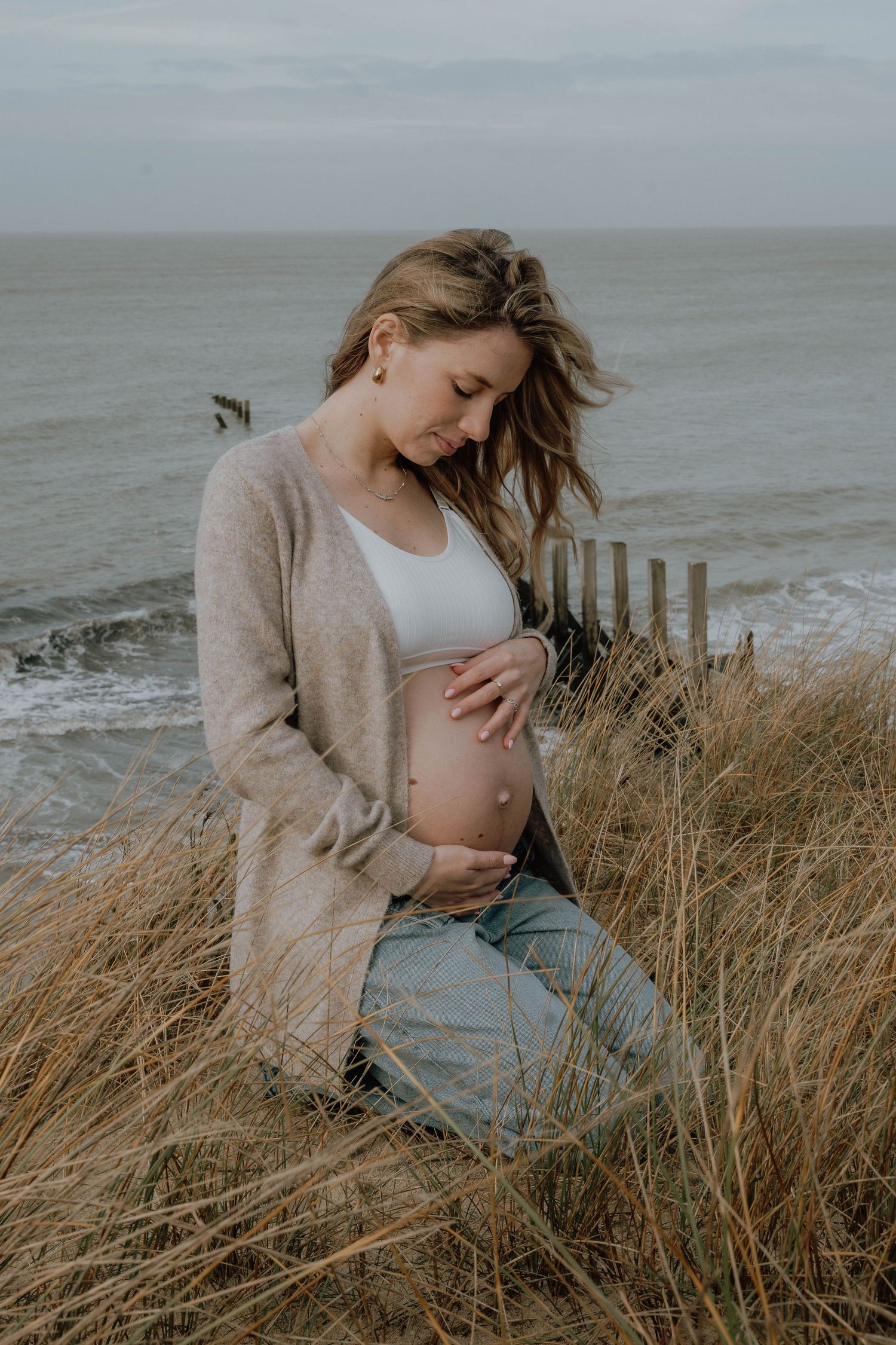 Pregnant woman cradling her baby bump while sitting in coastal dunes, wearing a beige cardigan and jeans.