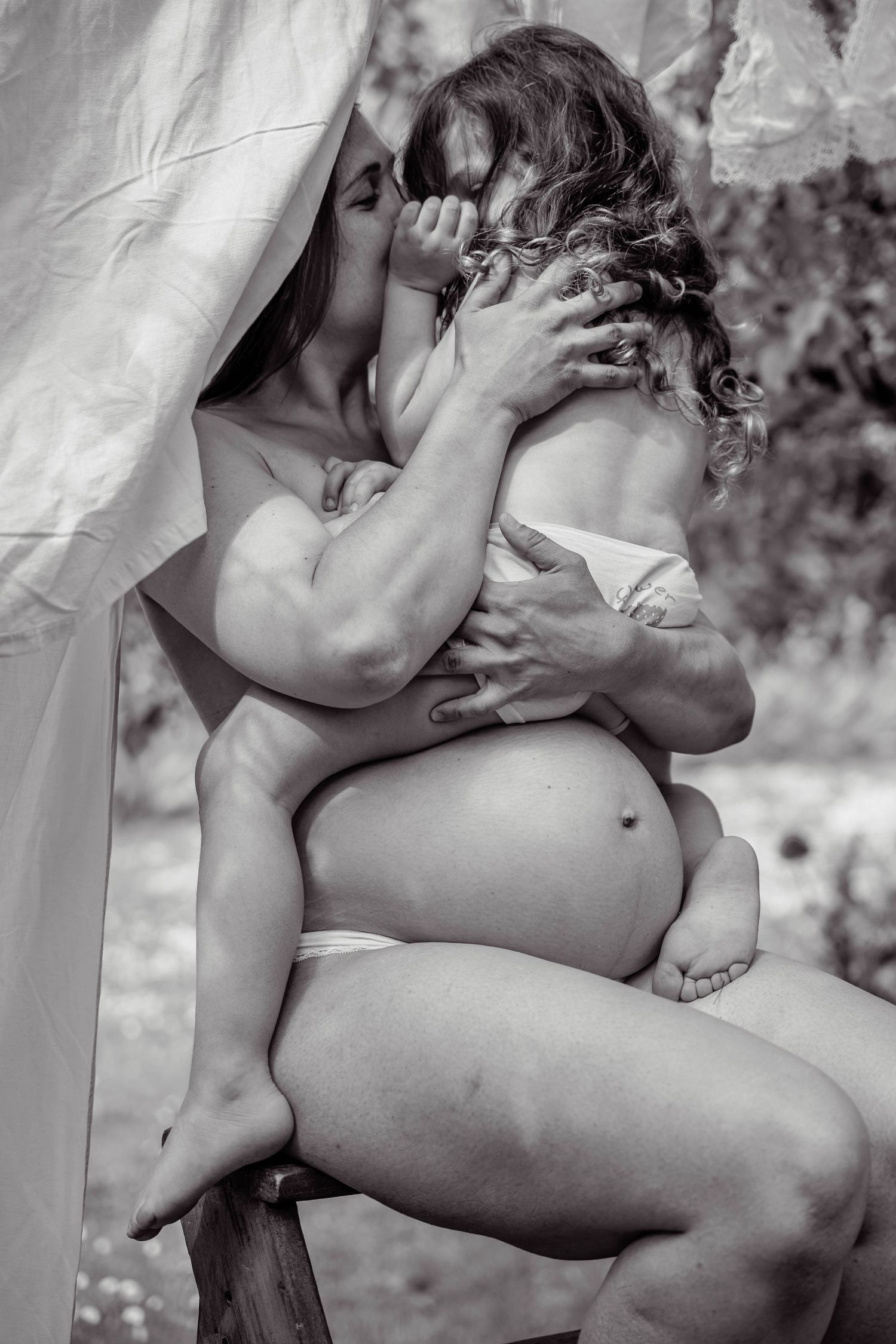 Black and white photo of a pregnant woman sitting outdoors, tenderly embracing and kissing a young child.