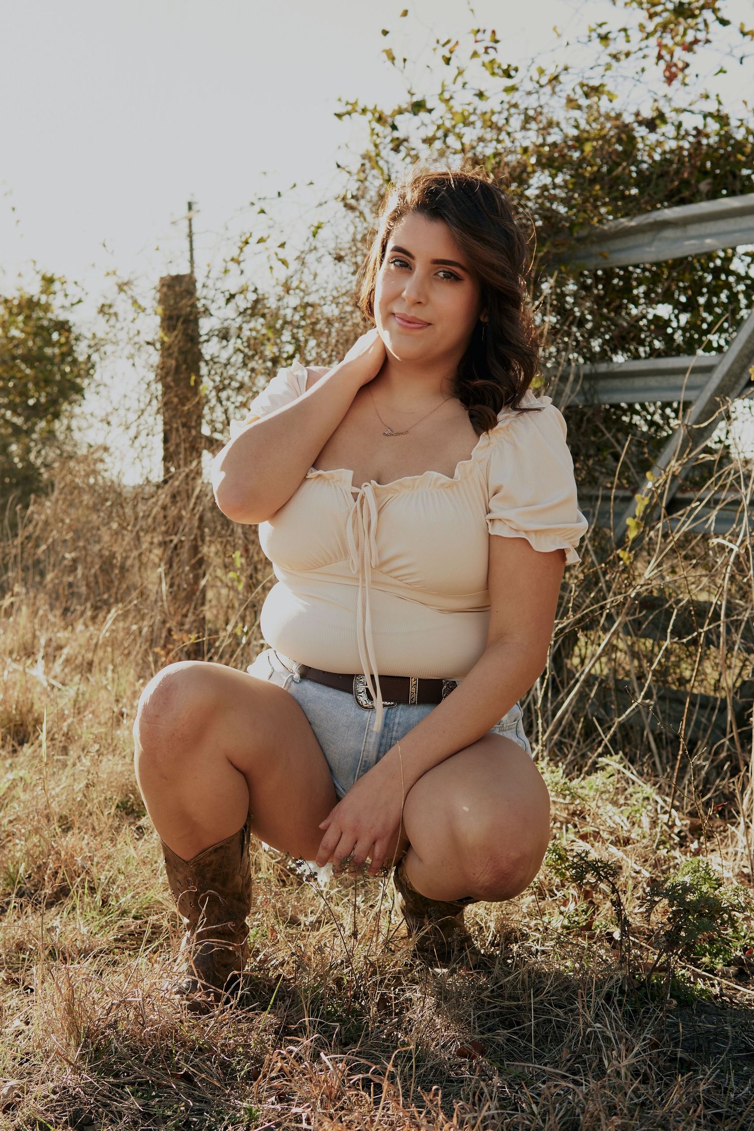 Woman in cream peasant top and denim shorts poses in rustic outdoor setting at sunset.