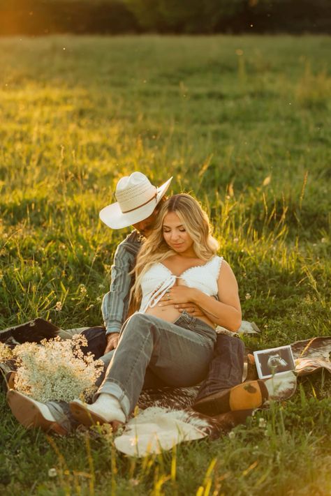 A couple sits together on a blanket in a sunlit meadow during golden hour with flowers and a sonogram photo nearby.