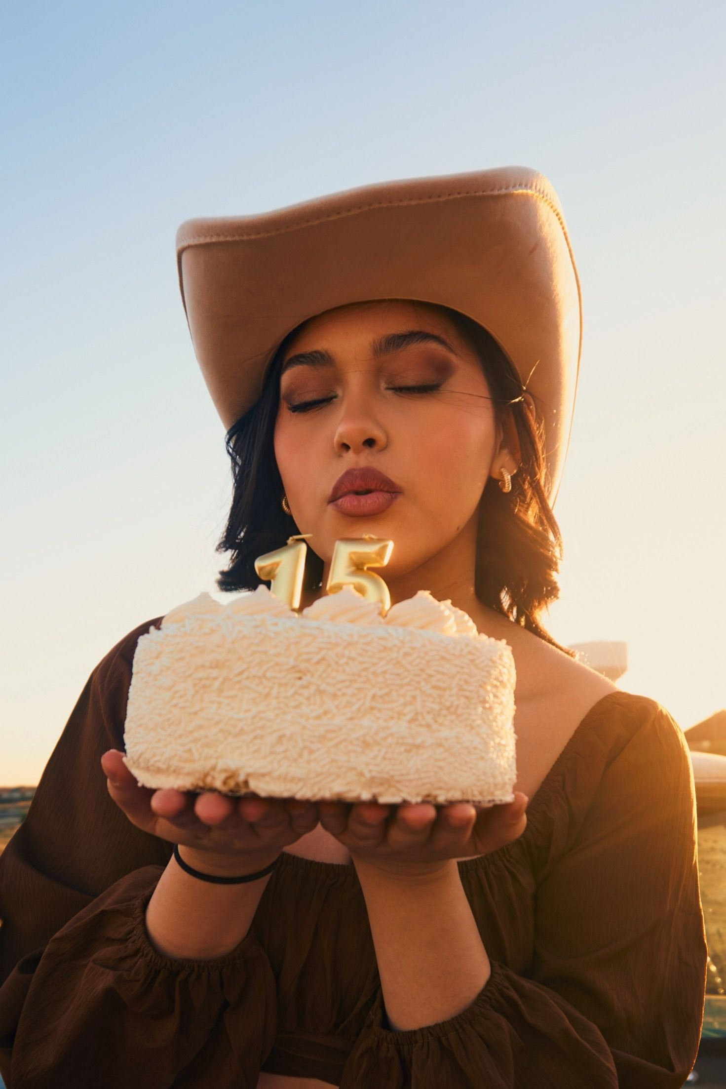 Person in cowboy hat holds white birthday cake with number 15 candles at sunset.
