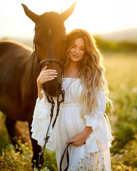 A brown horse and woman in a flowing white dress stand together in a golden sunlit meadow with wildflowers.