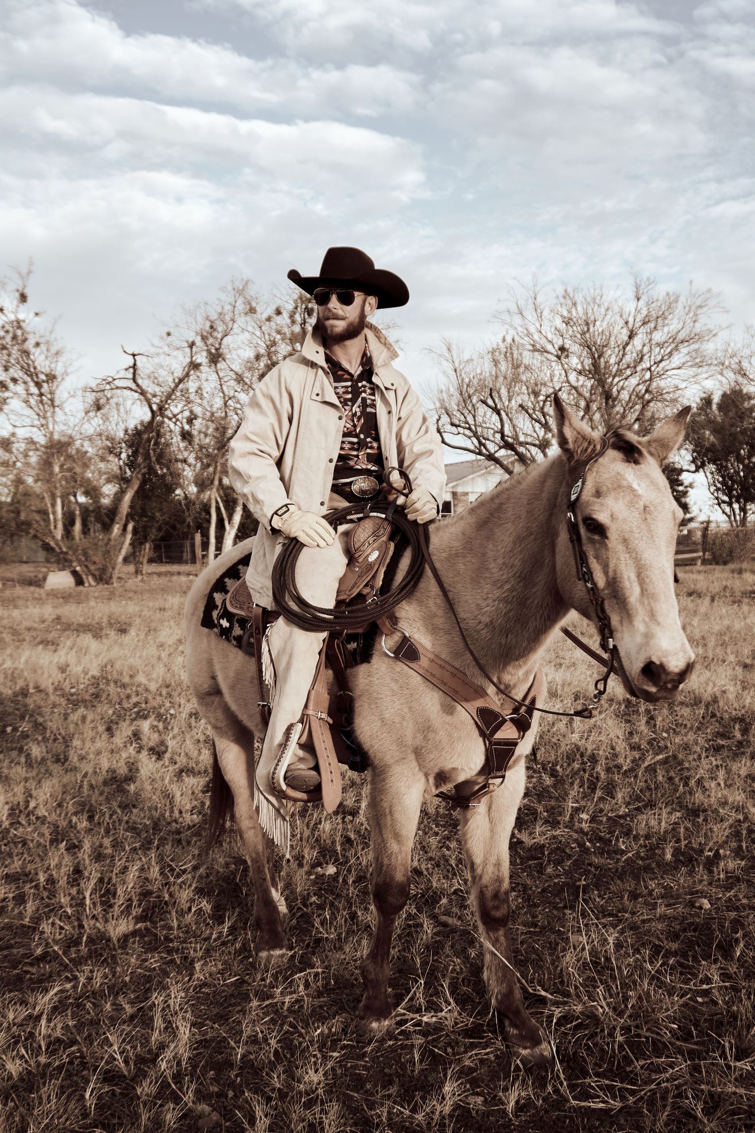 A sepia photograph showing a cowboy on horseback in a rural setting with bare trees.
