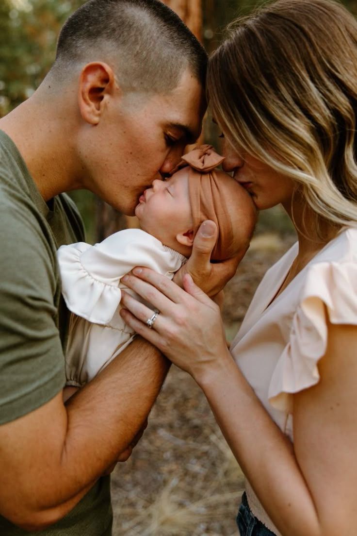 A loving couple shares a tender moment kissing their baby's head in an intimate outdoor family portrait.