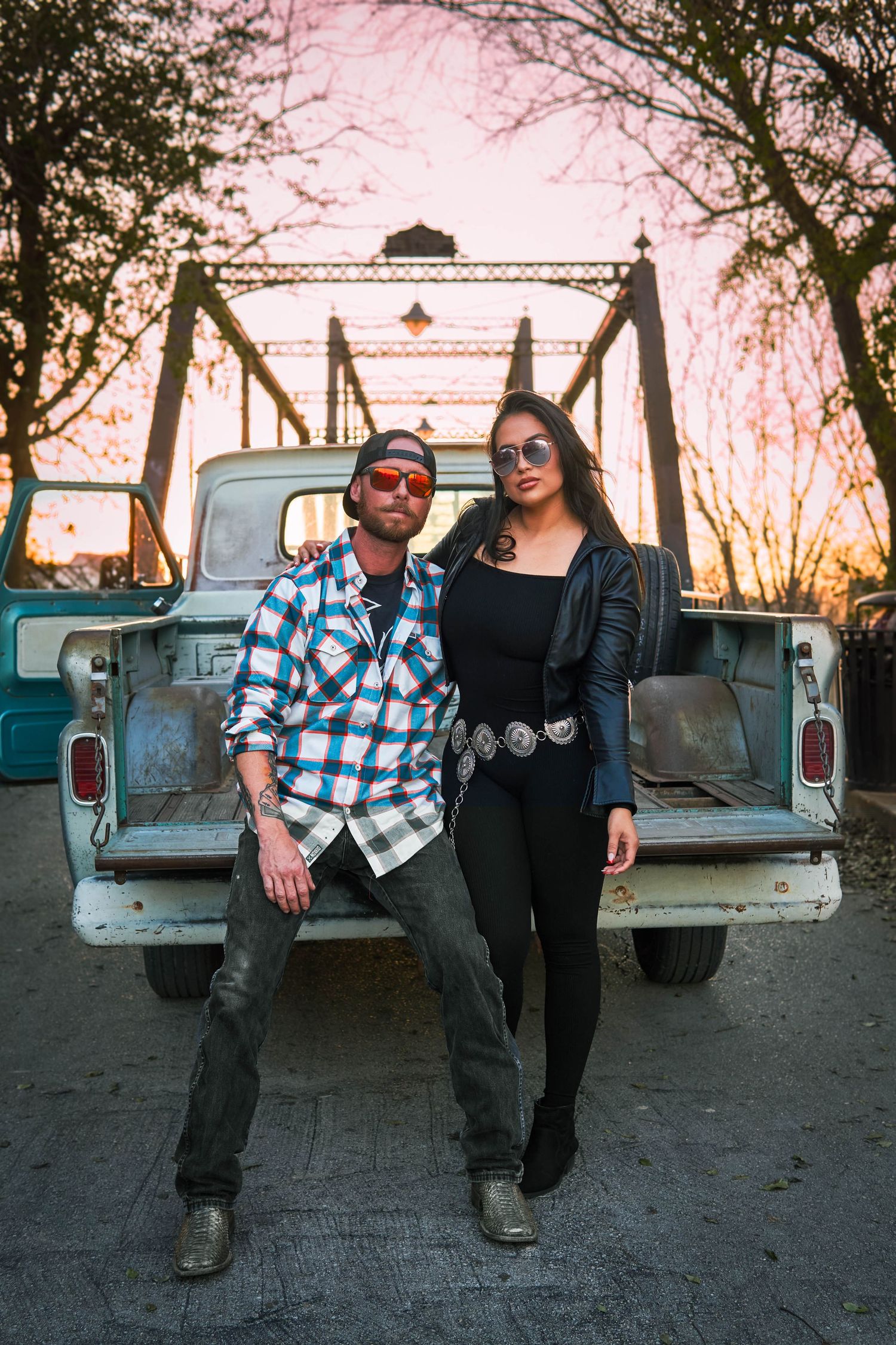 Two people stand together by a classic pickup truck with a metal bridge in the sunset background.