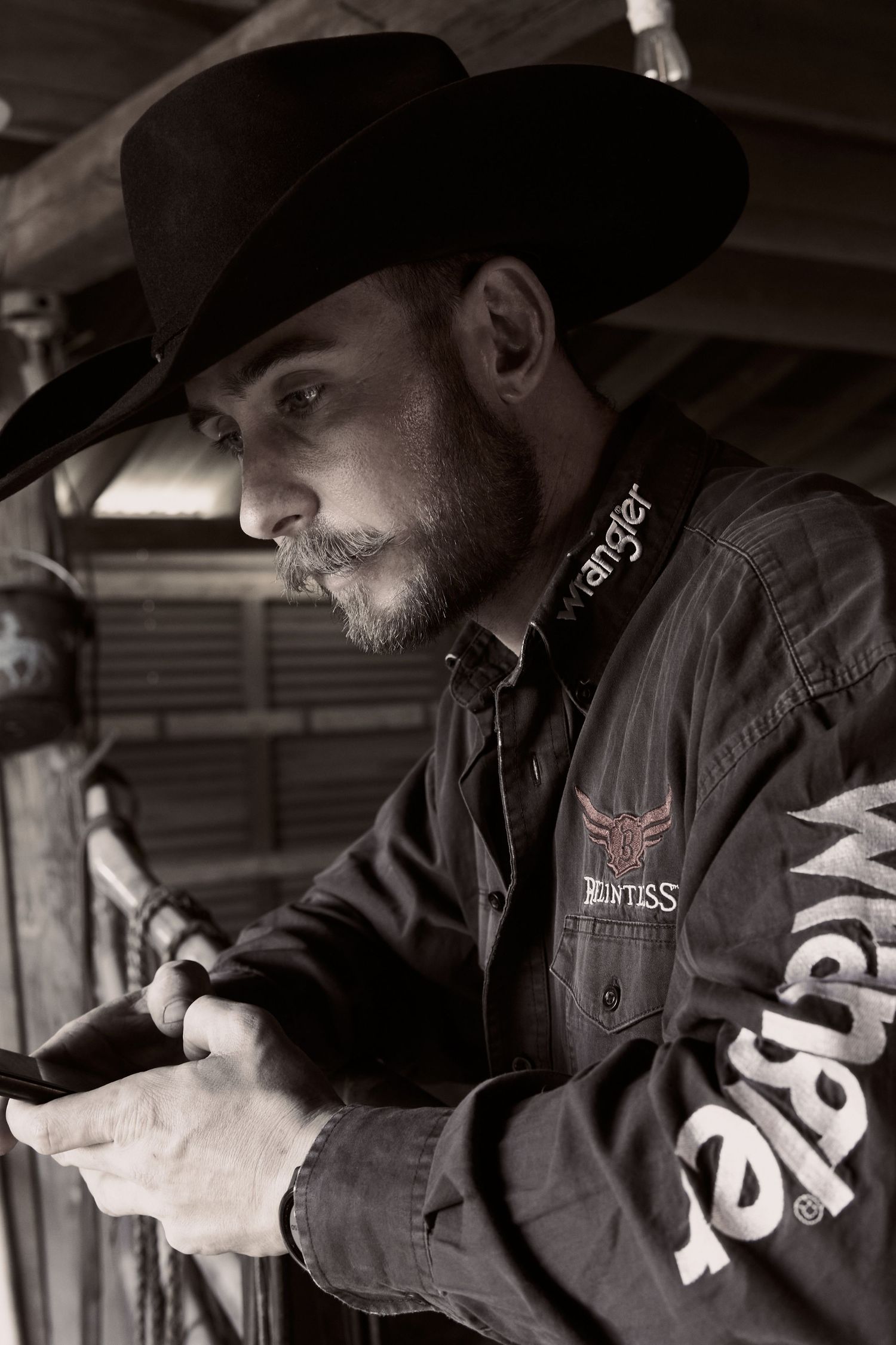 Side profile of cowboy in black hat looking at phone in dimly lit setting.