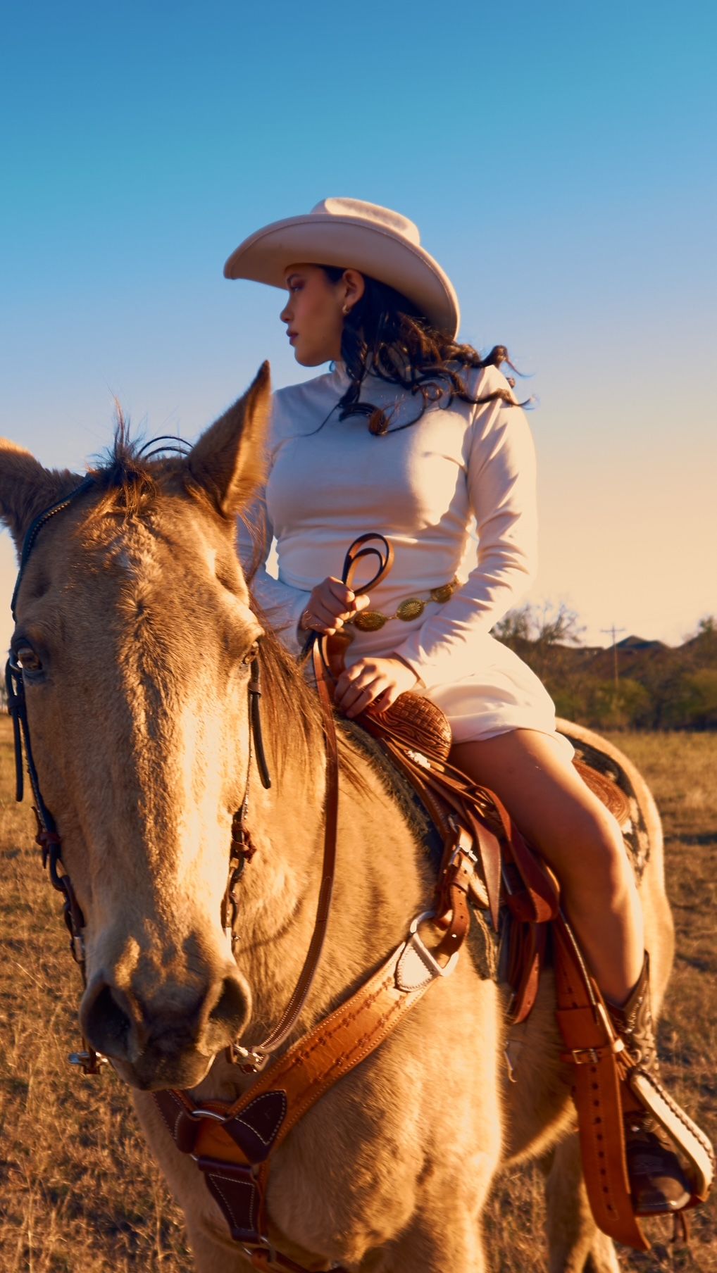 A rider in white western attire sits atop a light-colored horse against a blue sky at sunset.