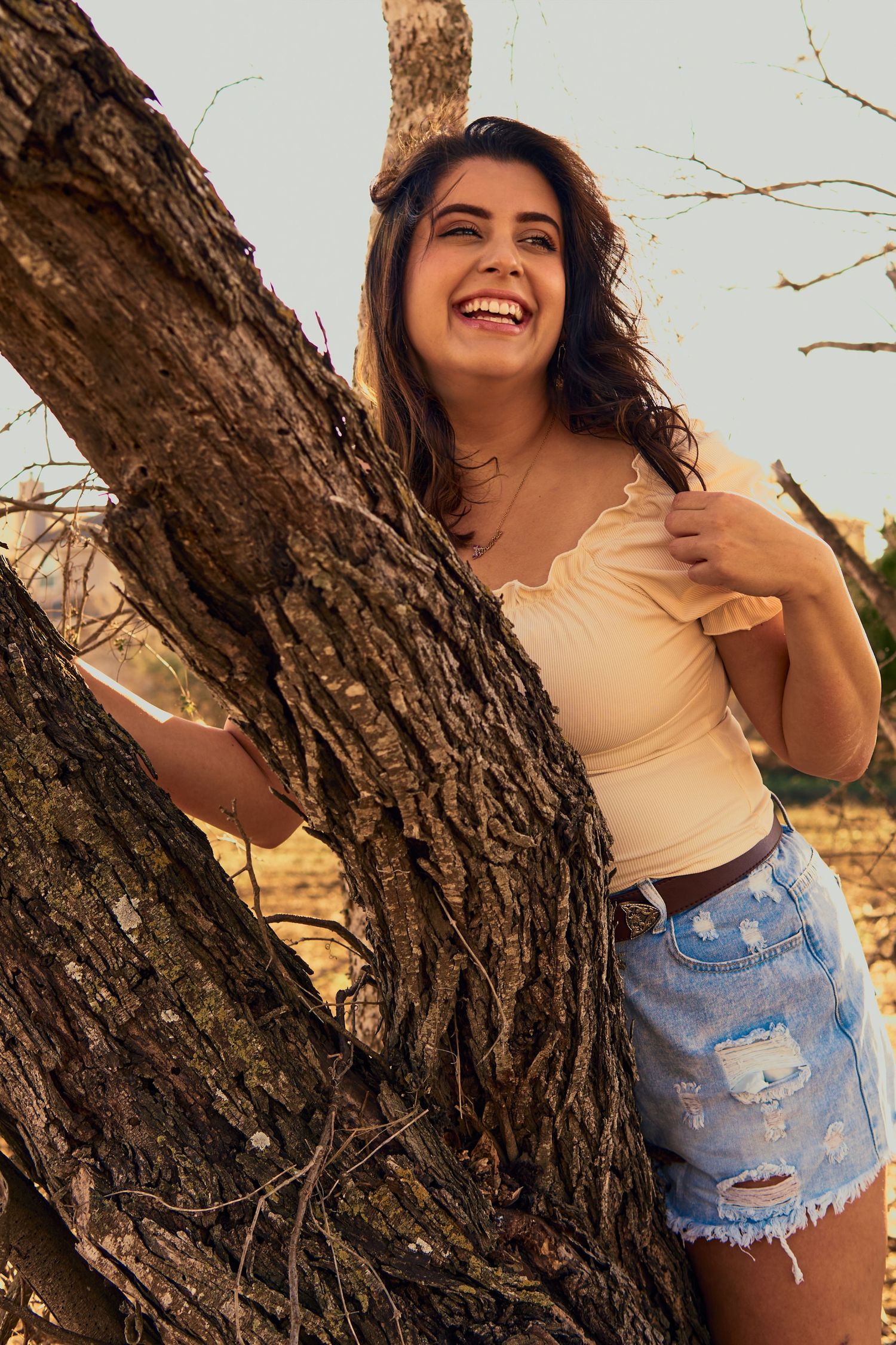 A woman in summer attire leans against a large tree trunk while smiling outdoors.