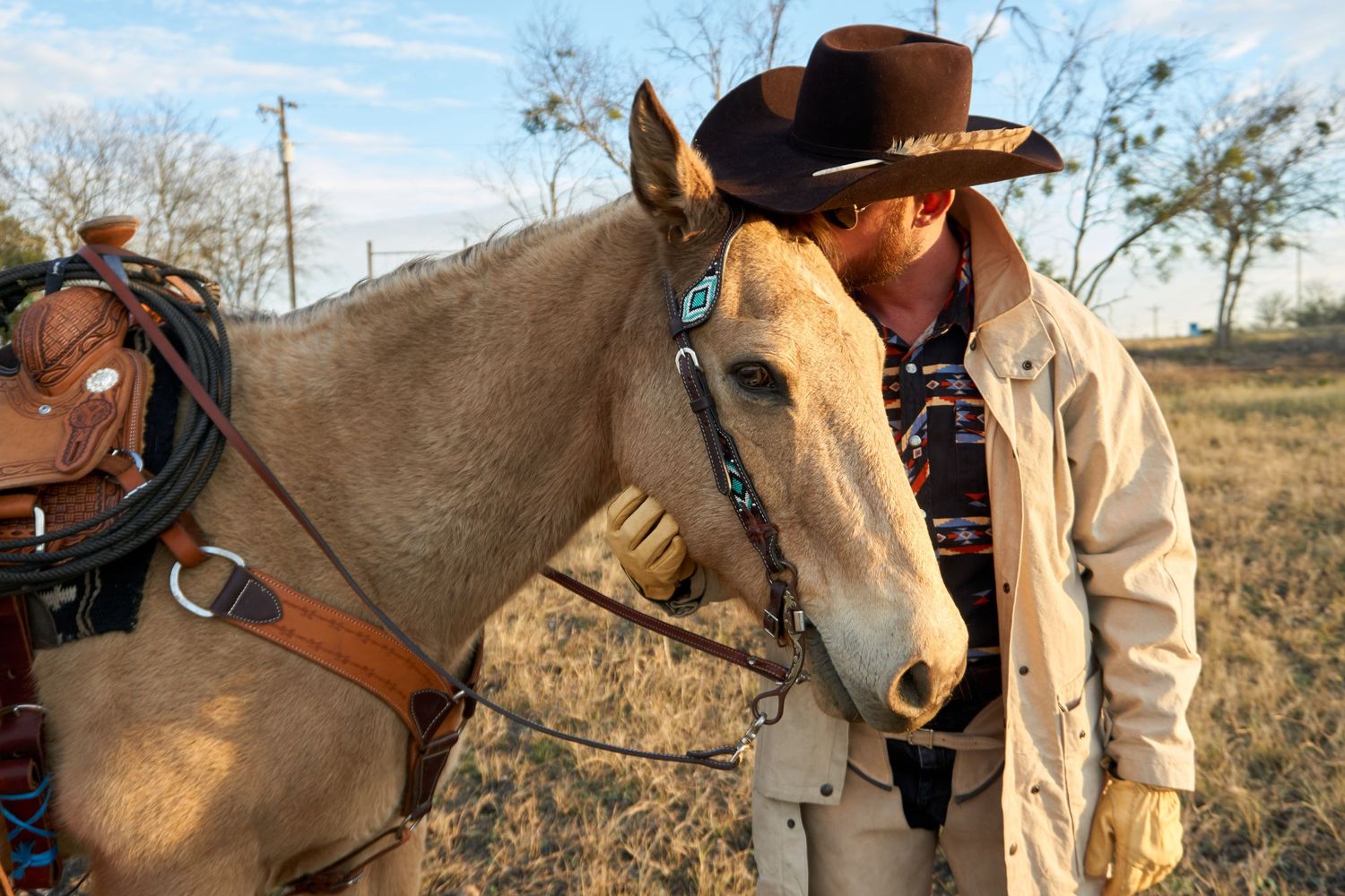 A light brown horse with leather tack stands outdoors as a cowboy in a brown hat and tan coat leans close.