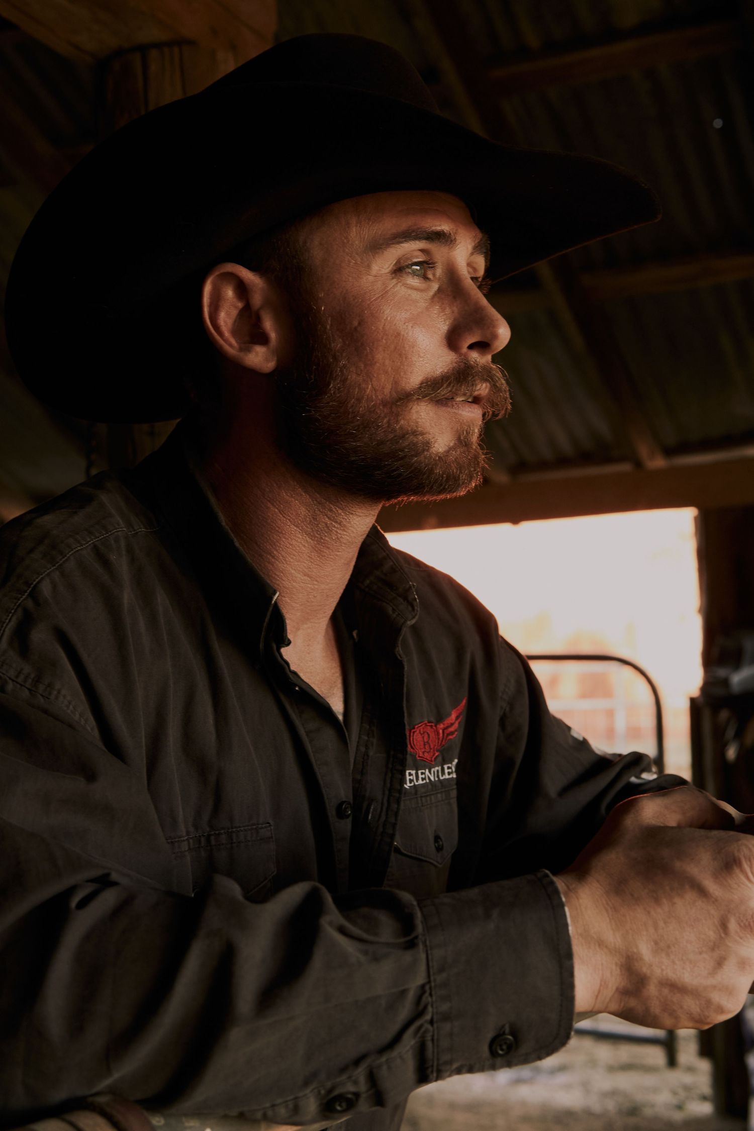 A cowboy in black hat and jacket gazes thoughtfully into the distance inside a barn.