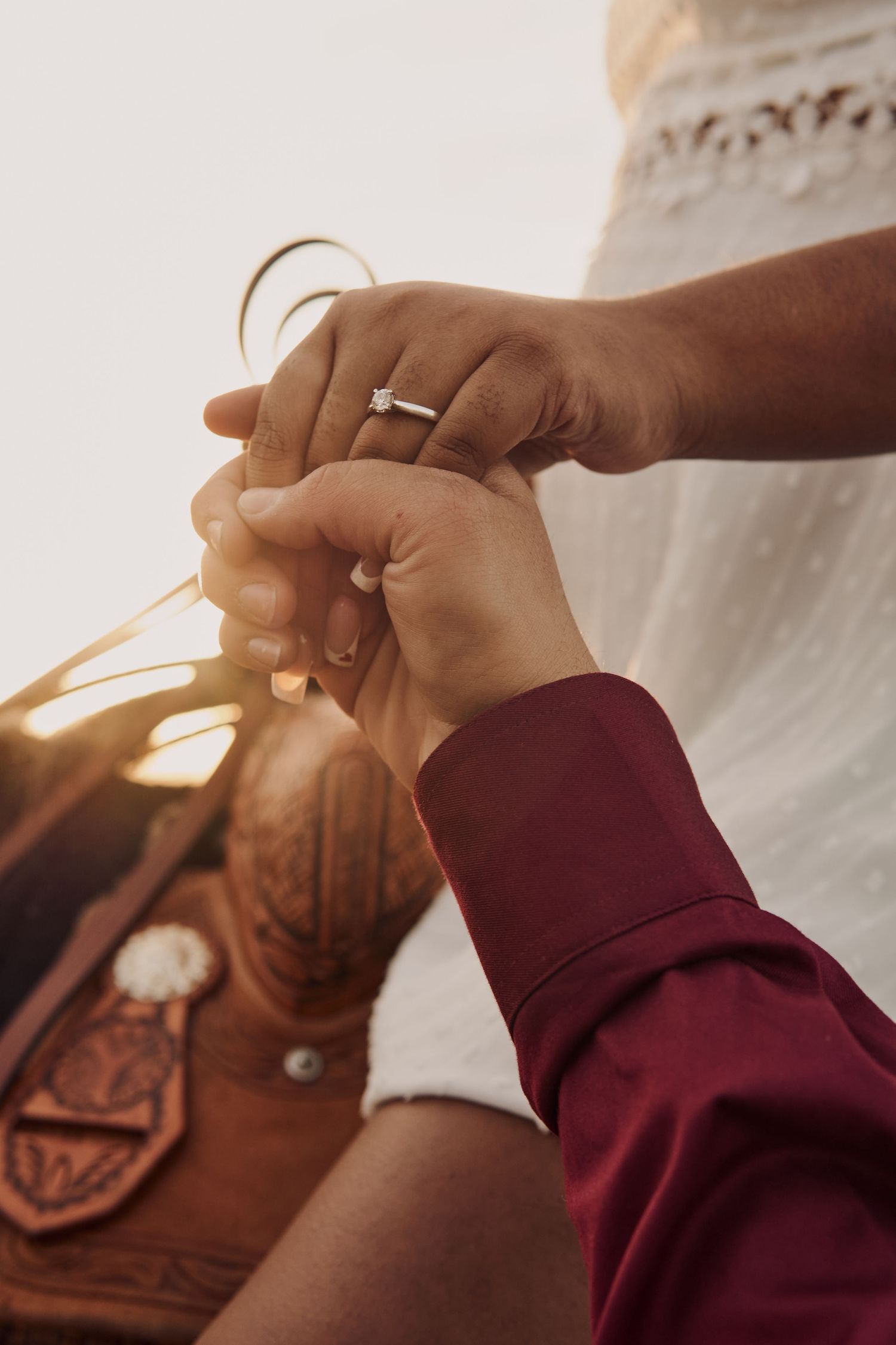 Close-up of hands with engagement ring exchanging string or thread in warm sunset light with leather glove visible.