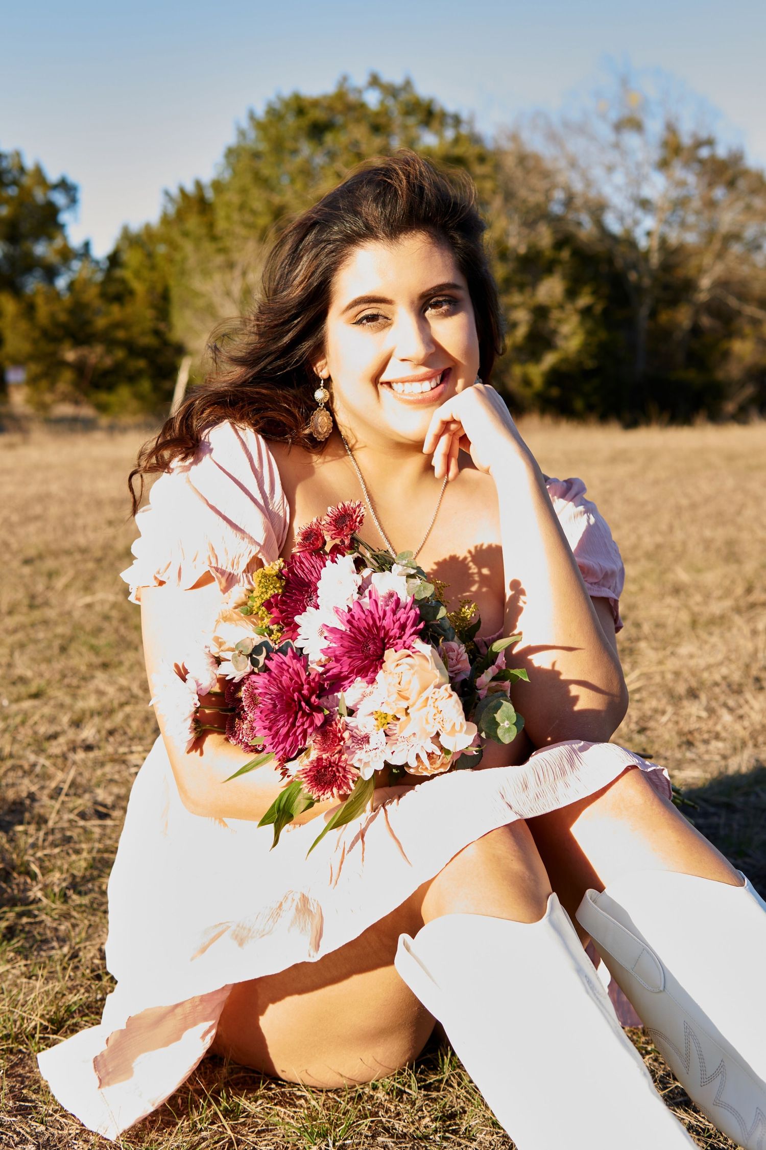 Portrait with pink floral bouquet sitting on white fence in sunny field.