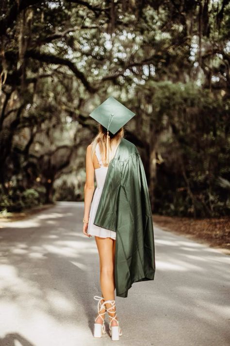 Ballet dancer in green graduation cap and gown poses en pointe on tree-lined path in sunlight.