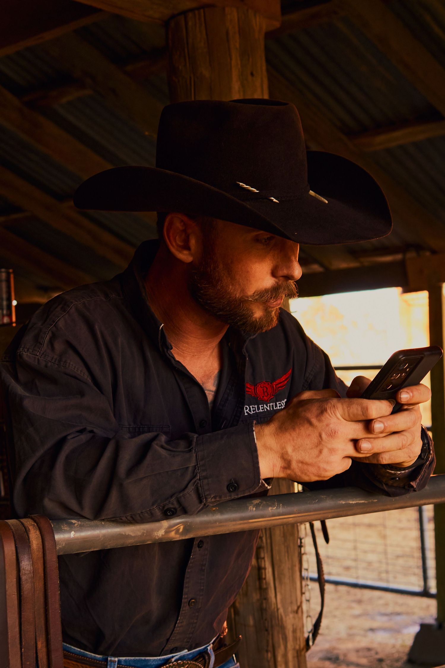 Cowboy in black hat using smartphone while leaning on wooden railing.
