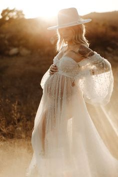 A romantic silhouette in a white flowing dress and wide brim hat stands against a golden sunset in a dreamy outdoor setting.