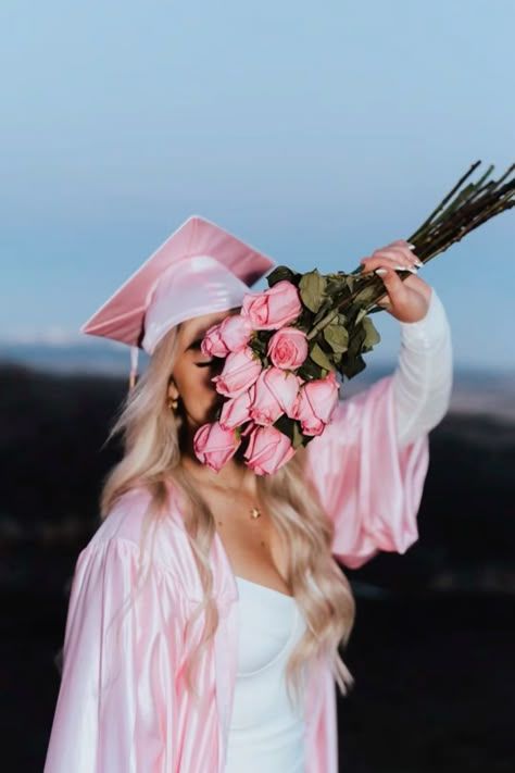 Woman in pink graduation gown holding pink roses against her face while posing outdoors at sunset.