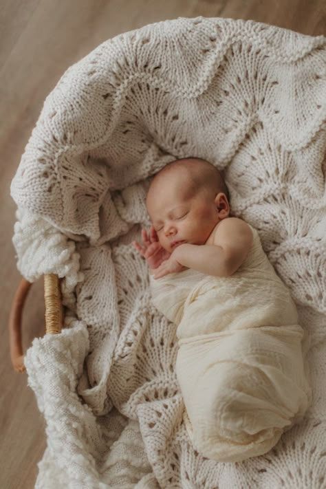 Sleeping newborn wrapped in cream fabric rests on knitted white blanket with fan pattern.