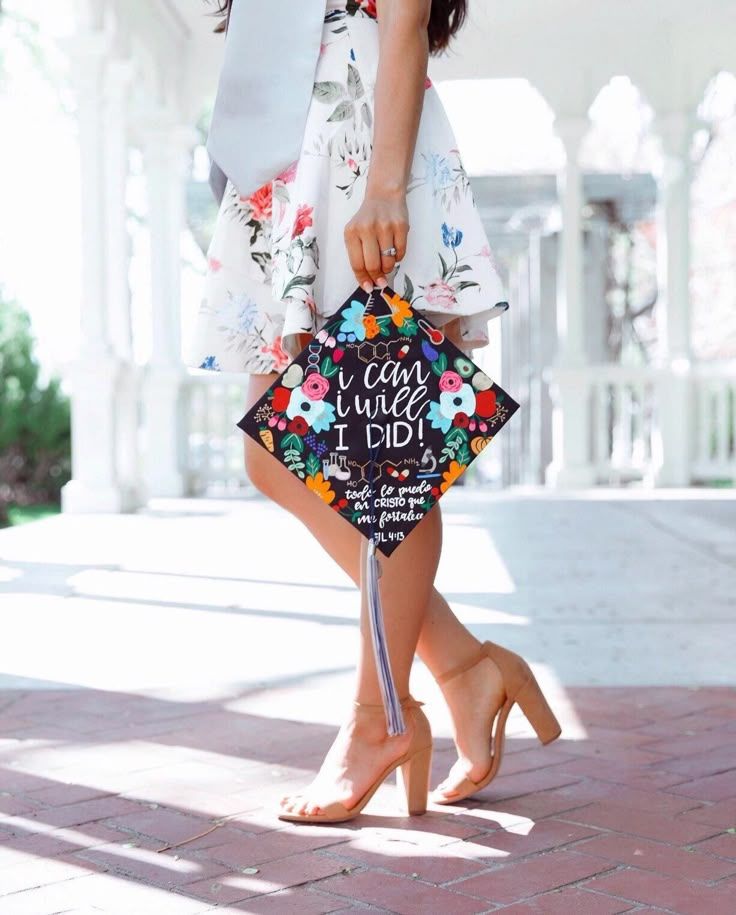 A close-up of a woman wearing a floral dress and nude heels, holding a decorated graduation cap near white columns.