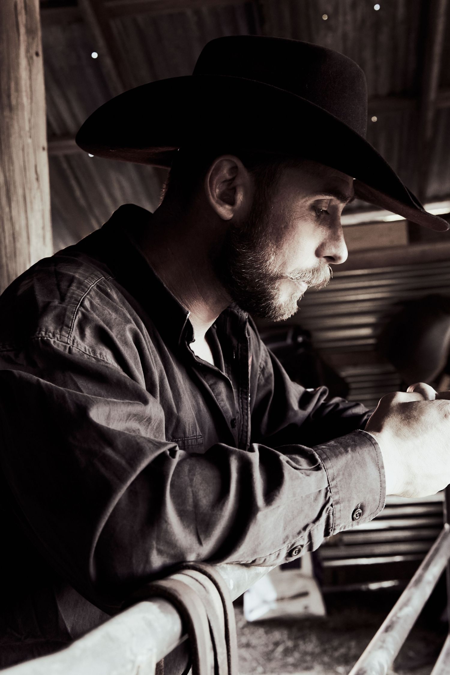 Dramatic black and white profile of rancher in cowboy hat leaning on fence.