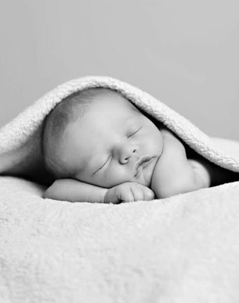 A sleeping newborn baby lies peacefully under a soft white blanket in a black and white photograph.