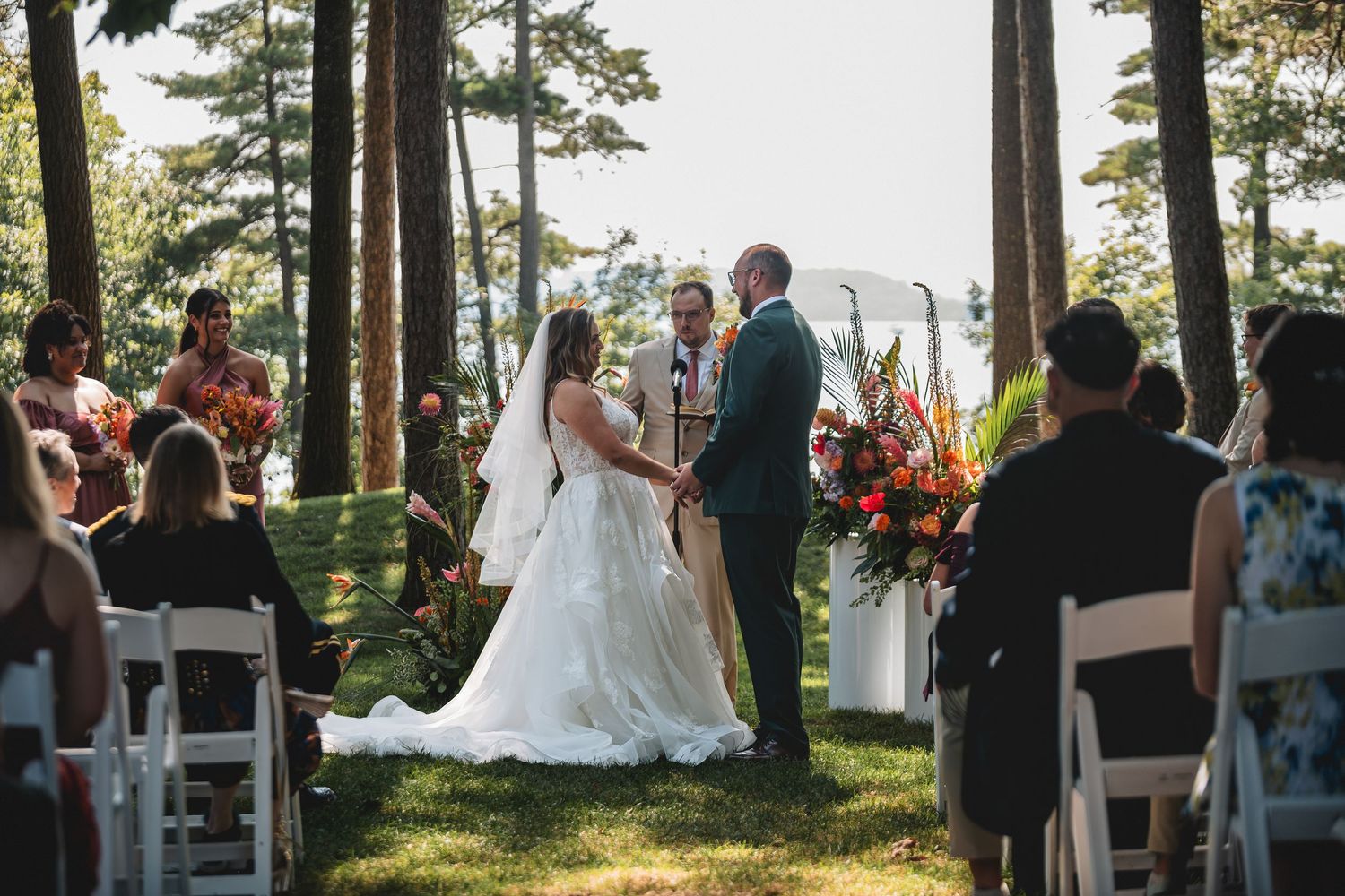 Wedding ceremony taking place outdoors among tall pine trees with guests seated in white chairs at sunset.