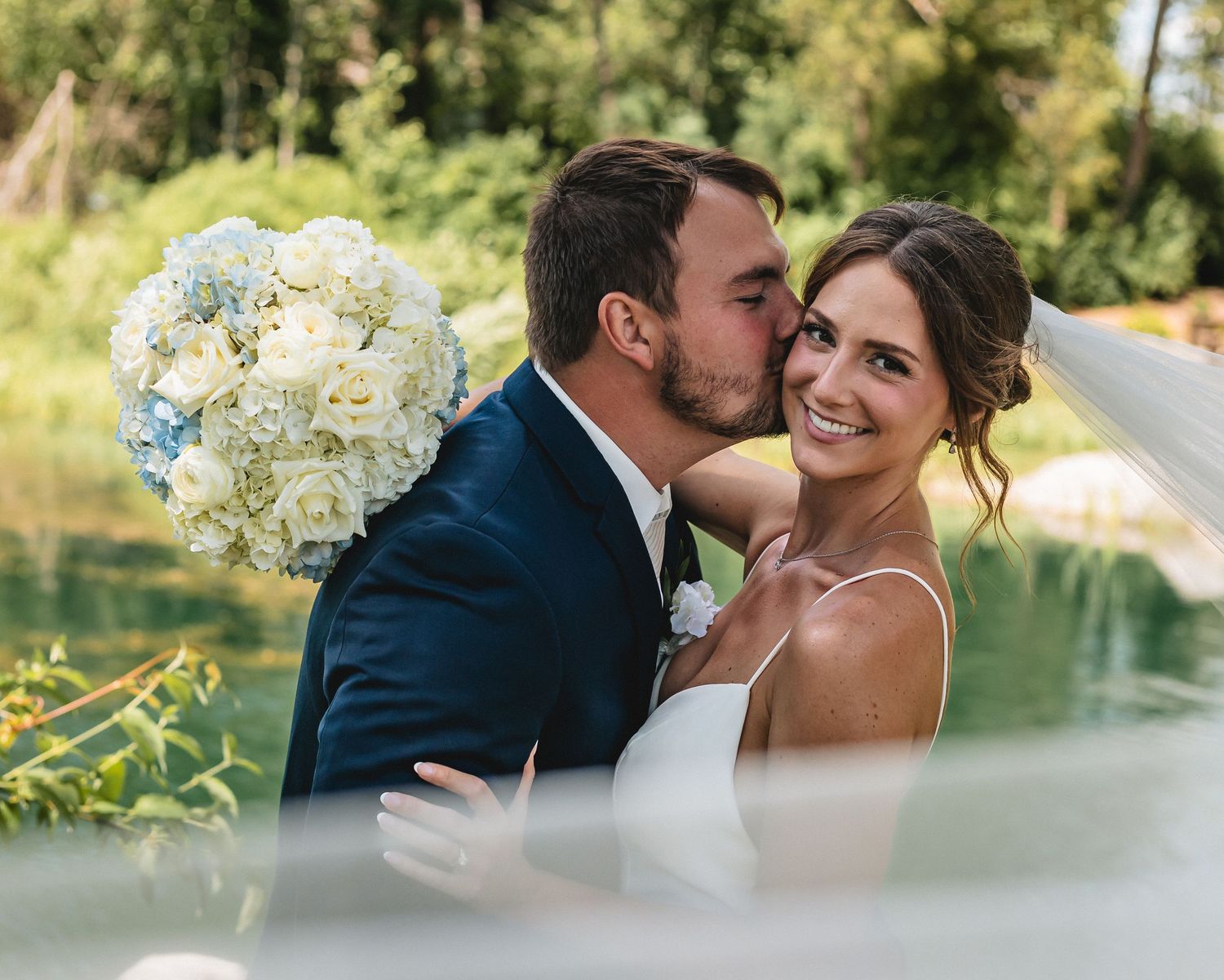 A groom in a navy suit kisses the cheek of a smiling bride holding a white and blue bouquet near a serene pond.