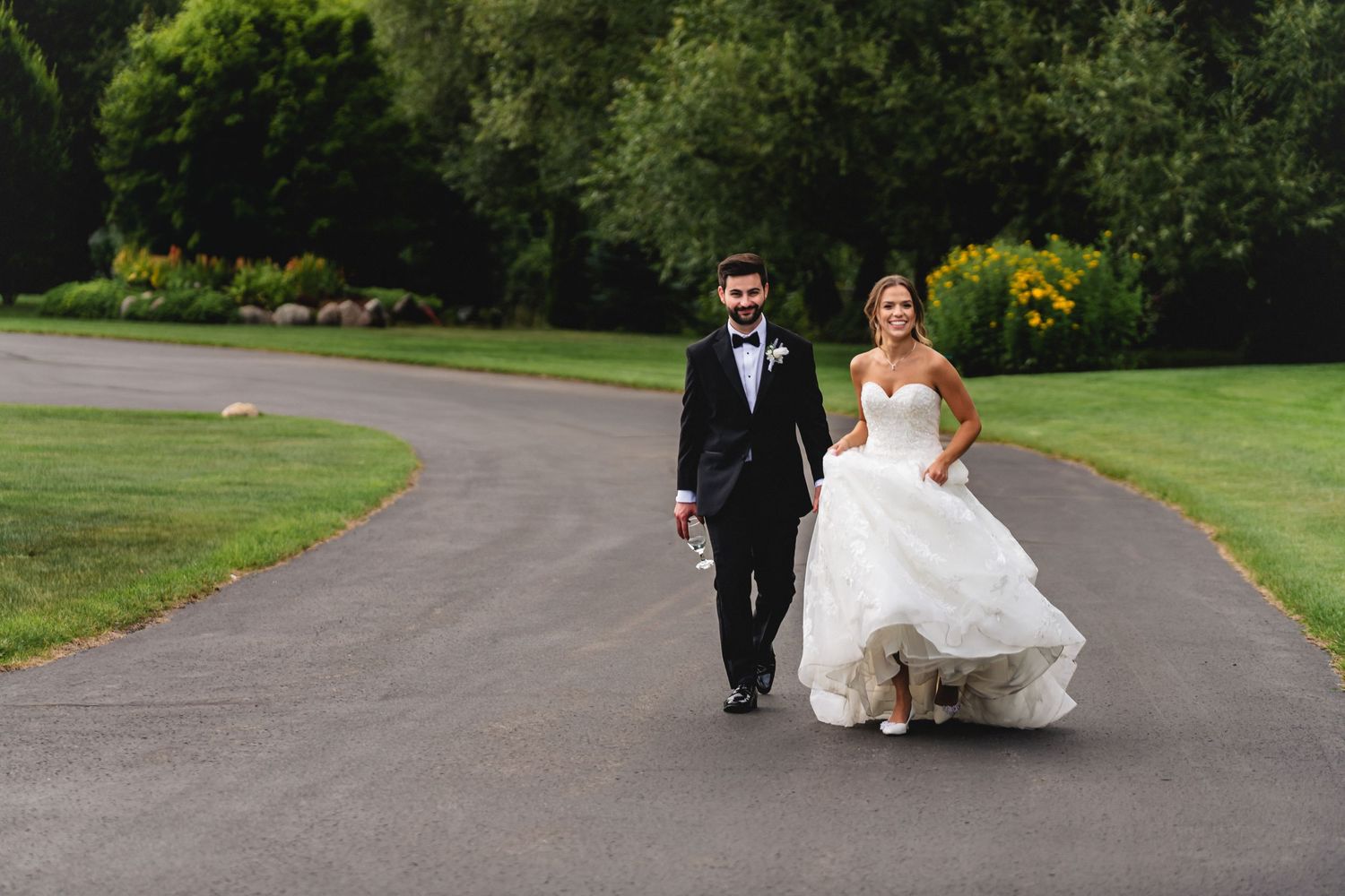 Bride and groom walking hand in hand on a tree-lined path, bride in a white ballgown, groom in a black tuxedo.