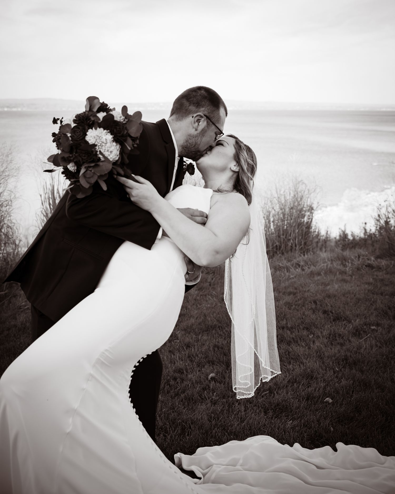 Black and white photo of a bride and groom sharing a romantic dip kiss outdoors with ocean views.