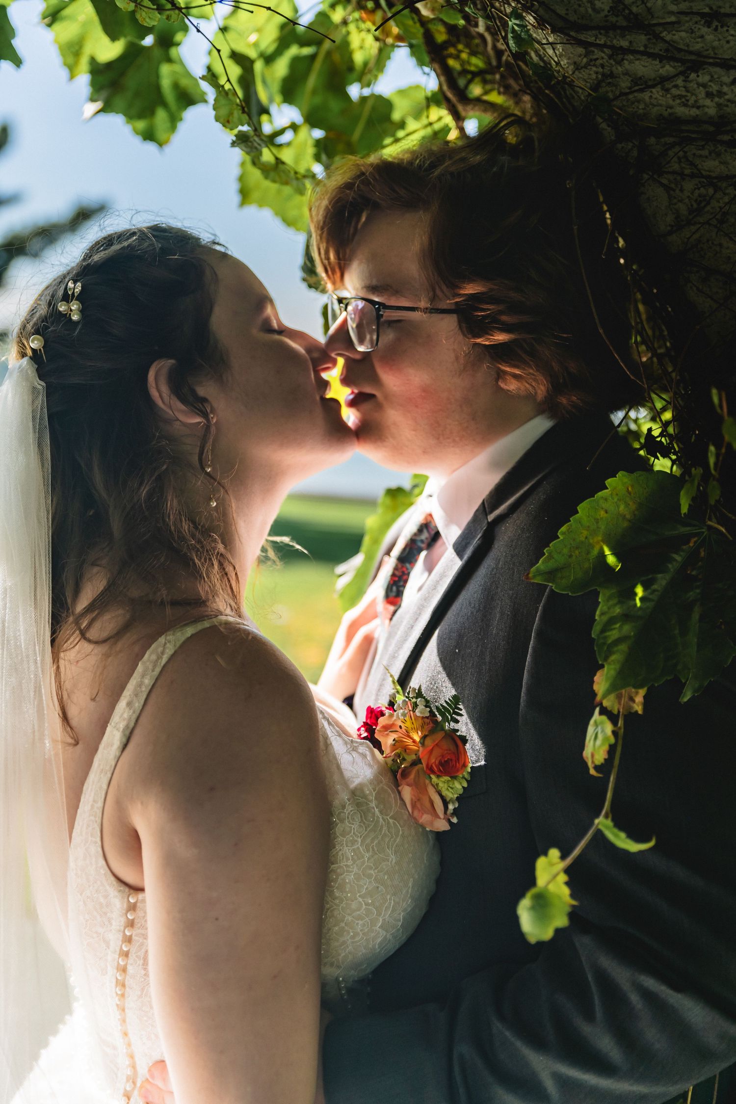 Bride and groom sharing a romantic kiss under a tree, with wedding veil and boutonniere visible.