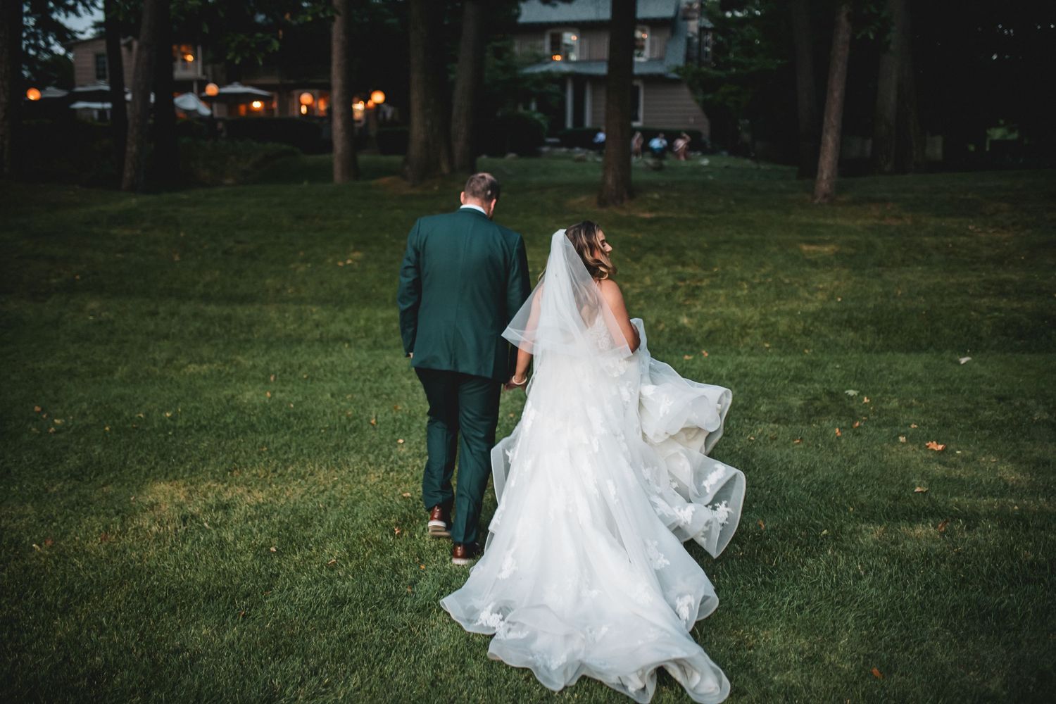 Newlywed couple walks hand in hand through a lush green garden during sunset, bride's white dress flowing in the breeze.