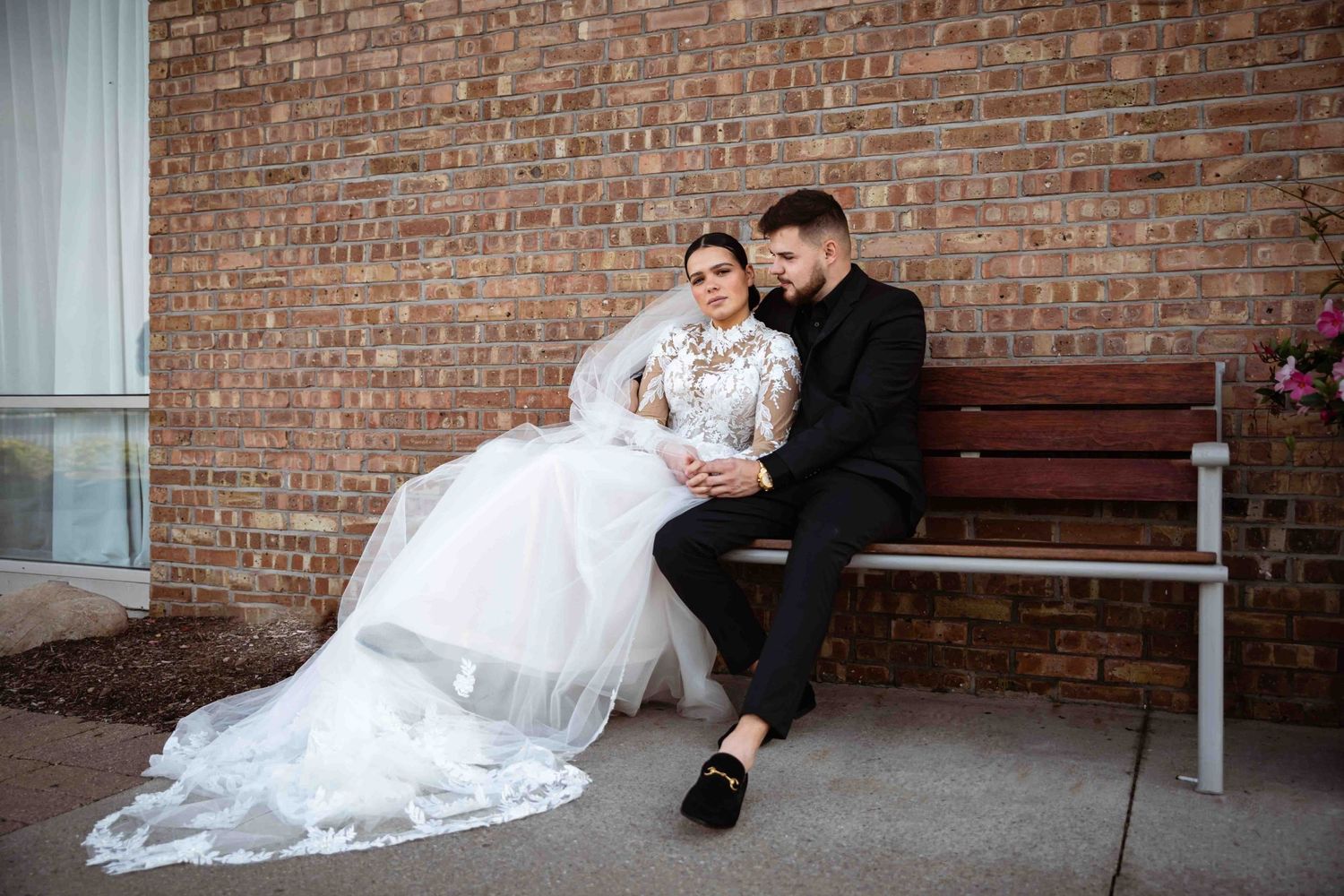 A couple in wedding attire sits together on a wooden bench against a rustic brick wall.