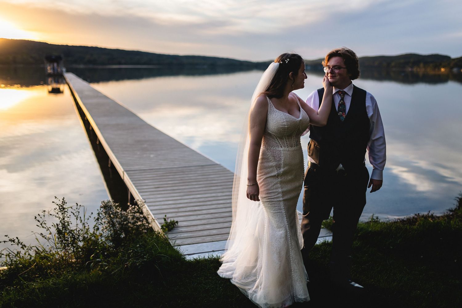 Newlywed couple walking along a wooden dock at sunset by a serene lake with mountains in the background.