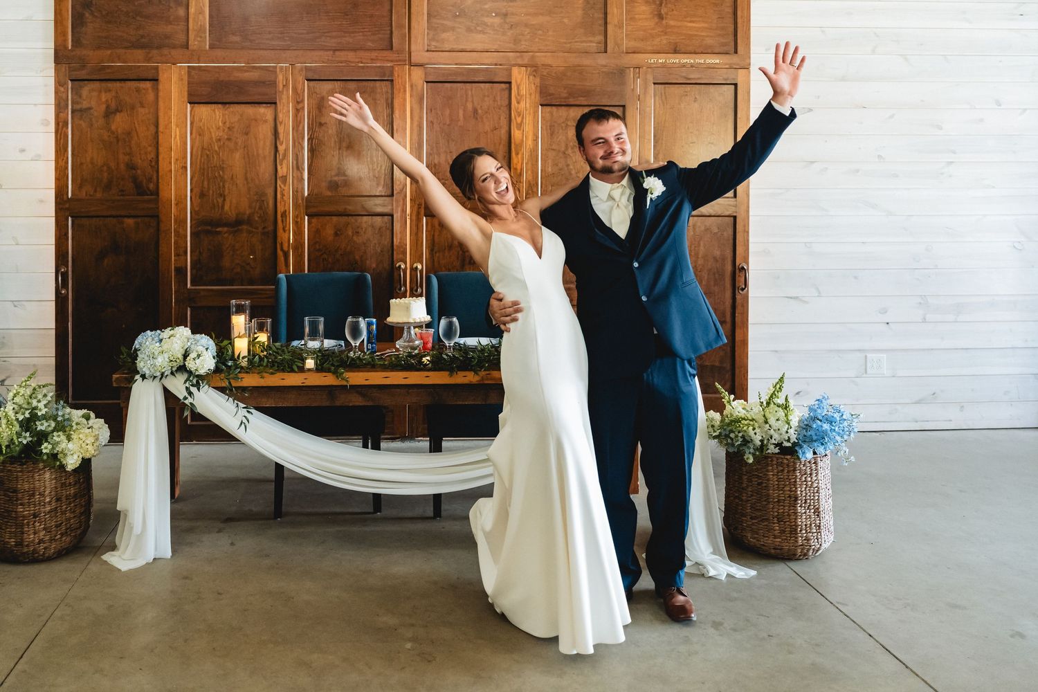 Joyful bride in white gown and groom in navy suit celebrating with raised arms at elegant wedding reception.
