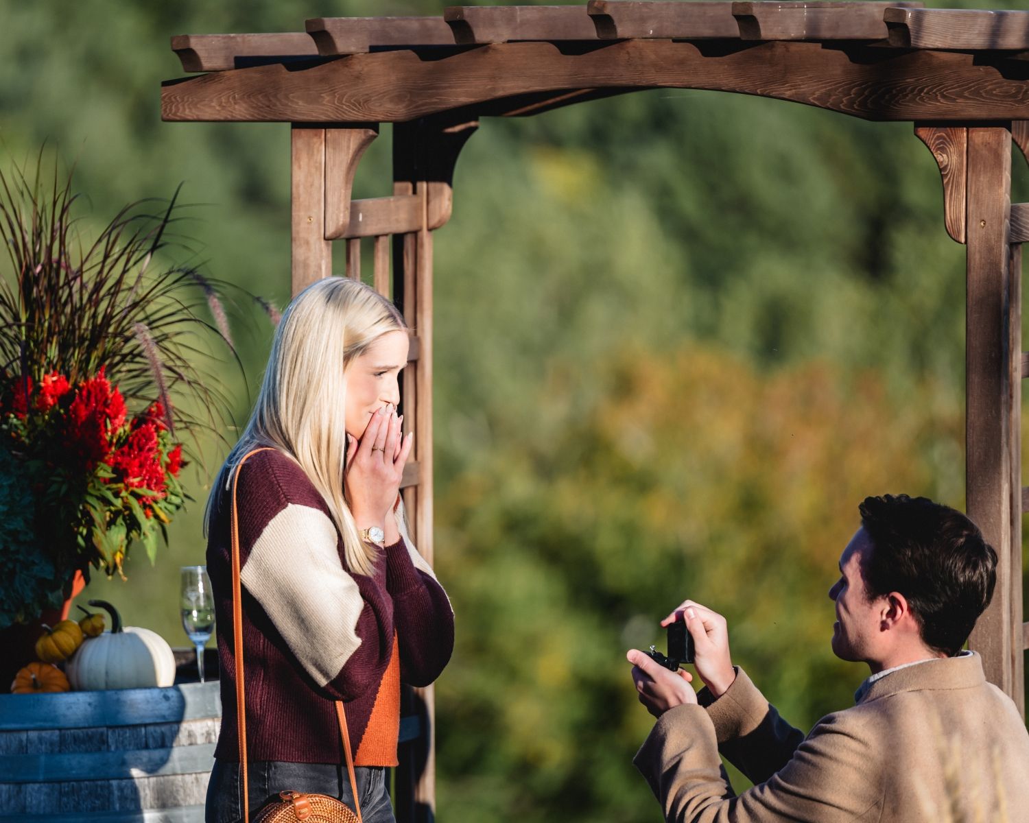 Man kneeling on one knee proposing to a surprised blonde woman under a wooden pergola in an autumn outdoor setting.