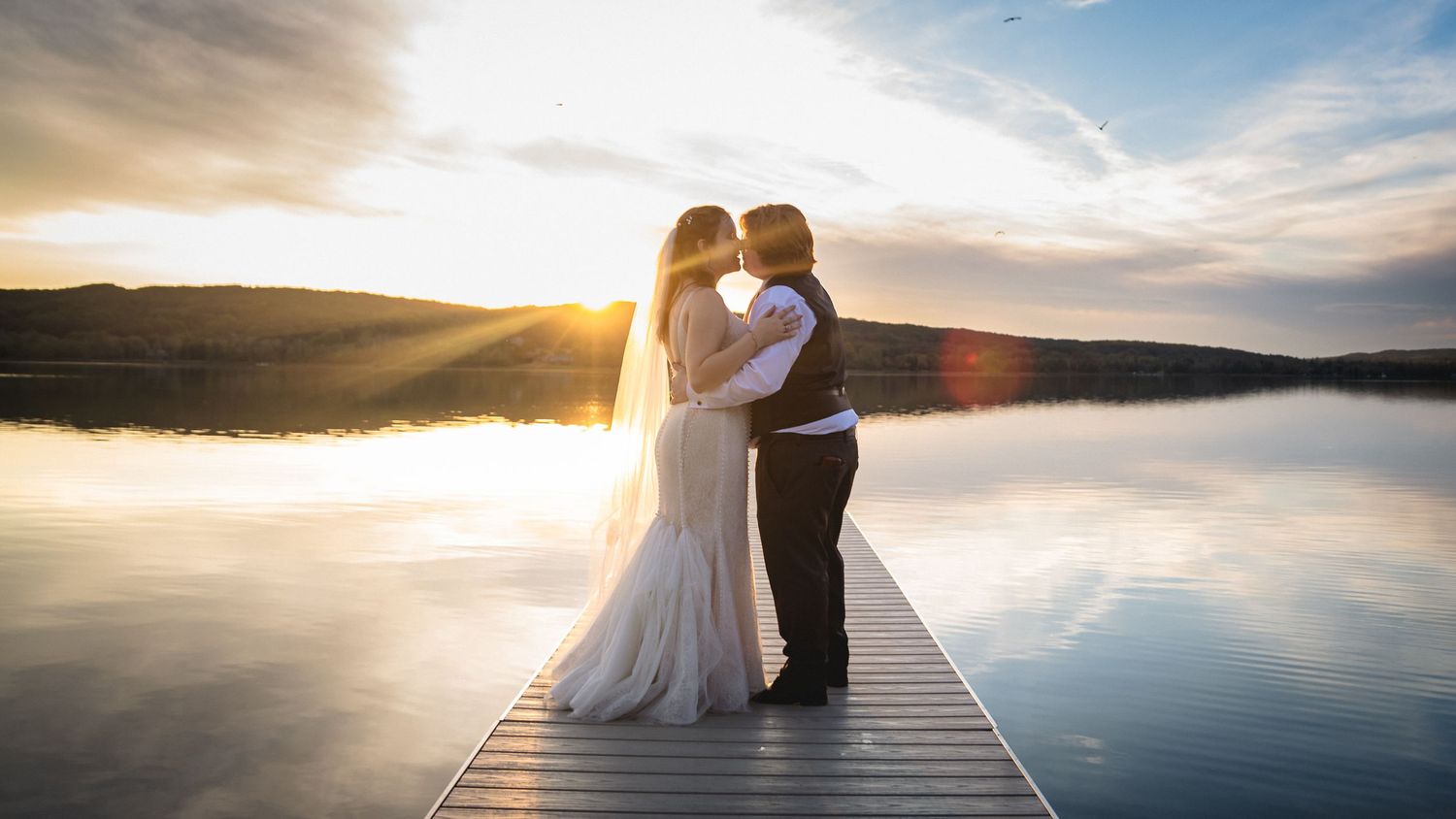 Bride and groom share a romantic kiss on a wooden dock at sunset over a calm reflective lake.