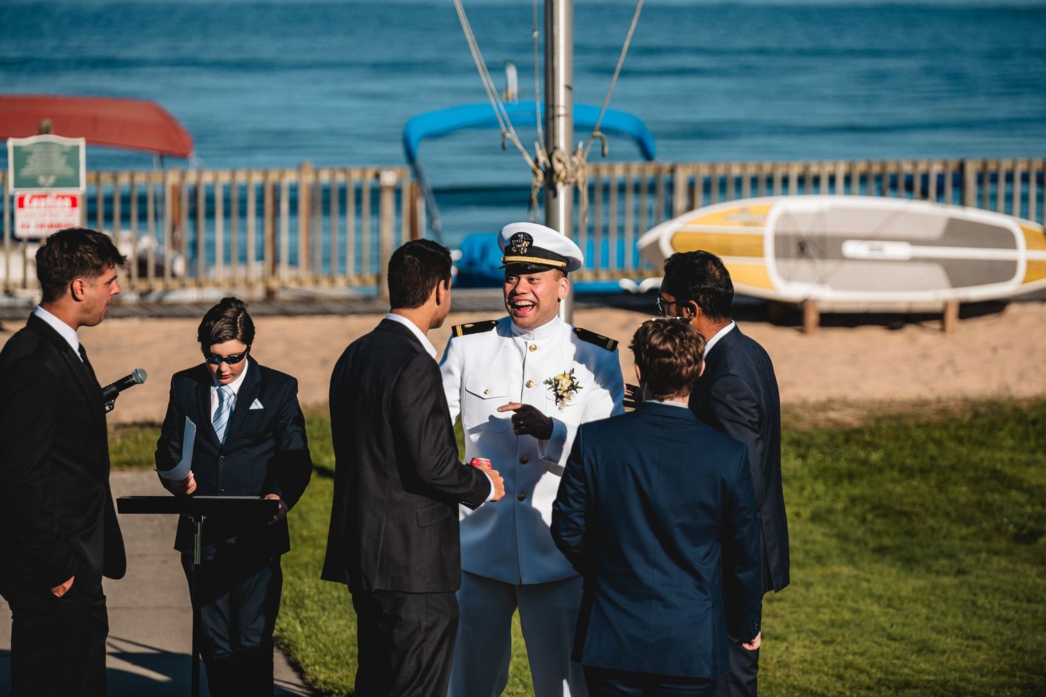 Naval officer in white uniform converses with suited guests near a sunny beachfront with colorful boats in background.