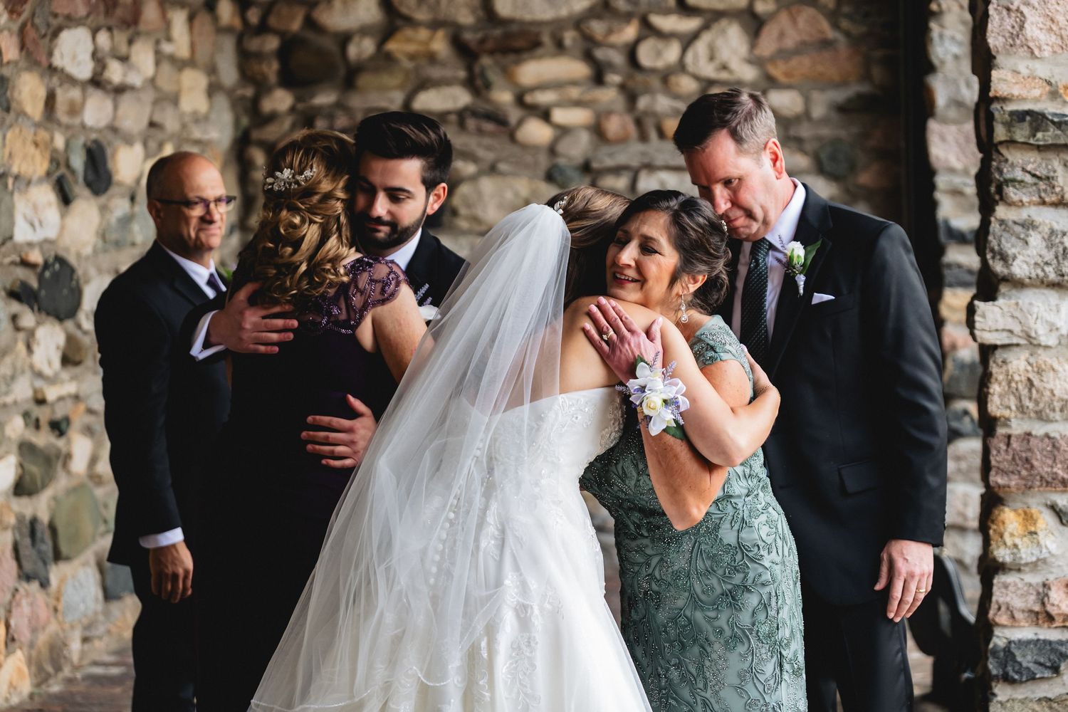 Bride and groom embracing family members in front of a stone wall at their wedding ceremony.