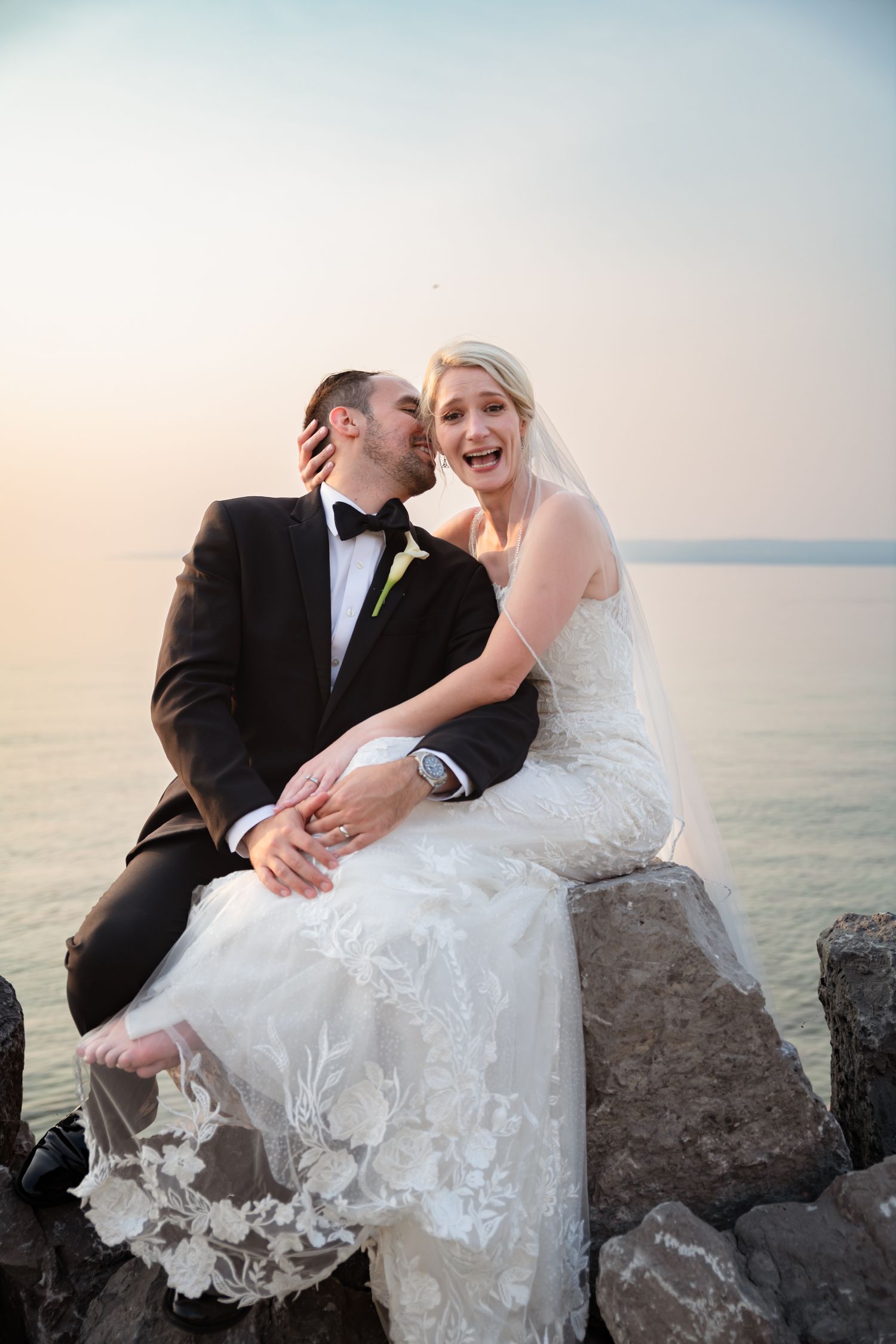 Bride and groom sitting on rocks by the water at sunset, laughing and sharing a kiss in wedding attire.