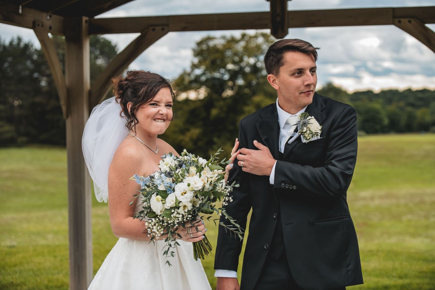 Bride and groom smiling under wooden pergola, bride holding white floral bouquet, groom in dark suit at outdoor wedding.