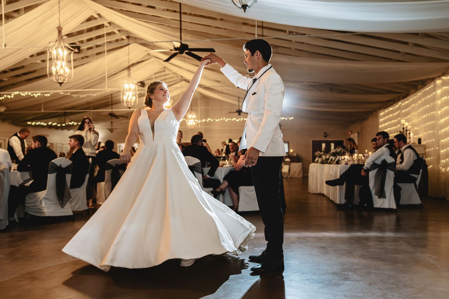 Bride and groom share their first dance in elegant wedding venue with string lights and guests seated nearby.