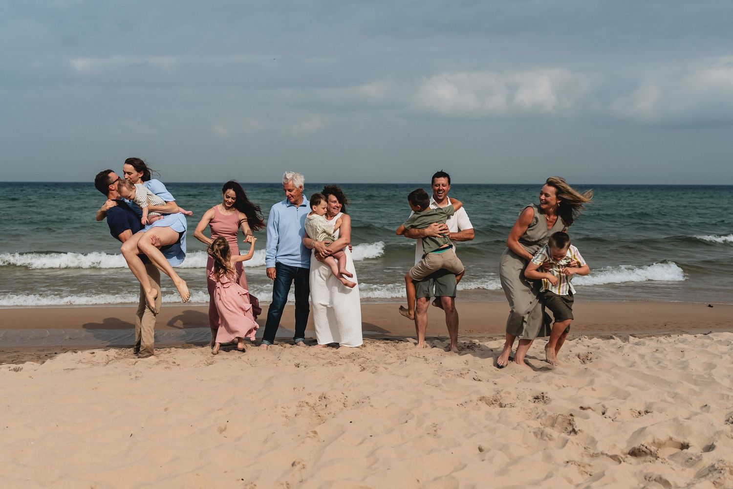 Multi-generational family laughing and playing together on a sandy beach with ocean waves in the background.