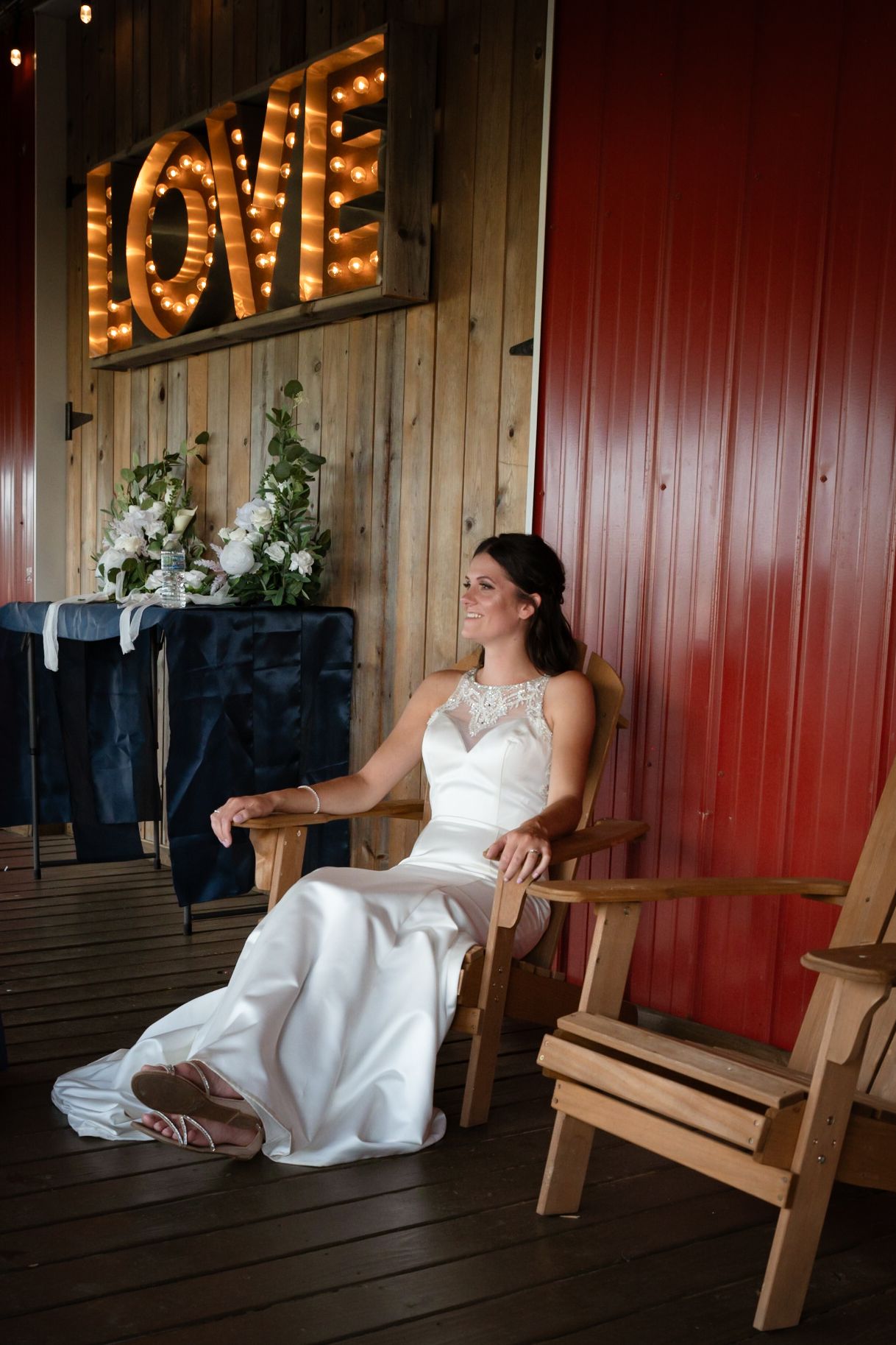 Bride in white gown sitting in wooden chair at rustic barn wedding venue with illuminated LOVE sign backdrop.