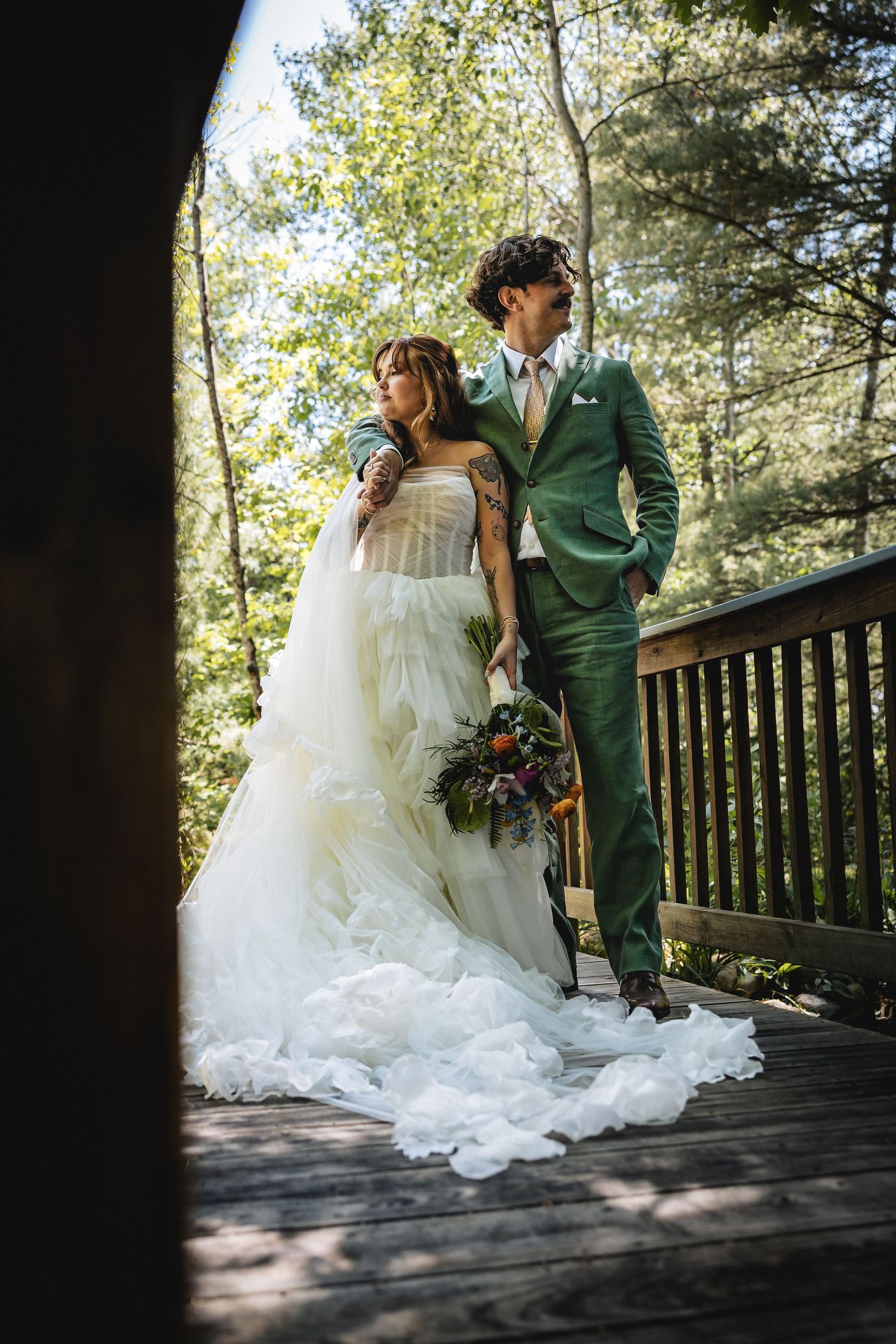 Bride in white tulle gown and groom in green suit pose on wooden deck surrounded by lush forest trees.