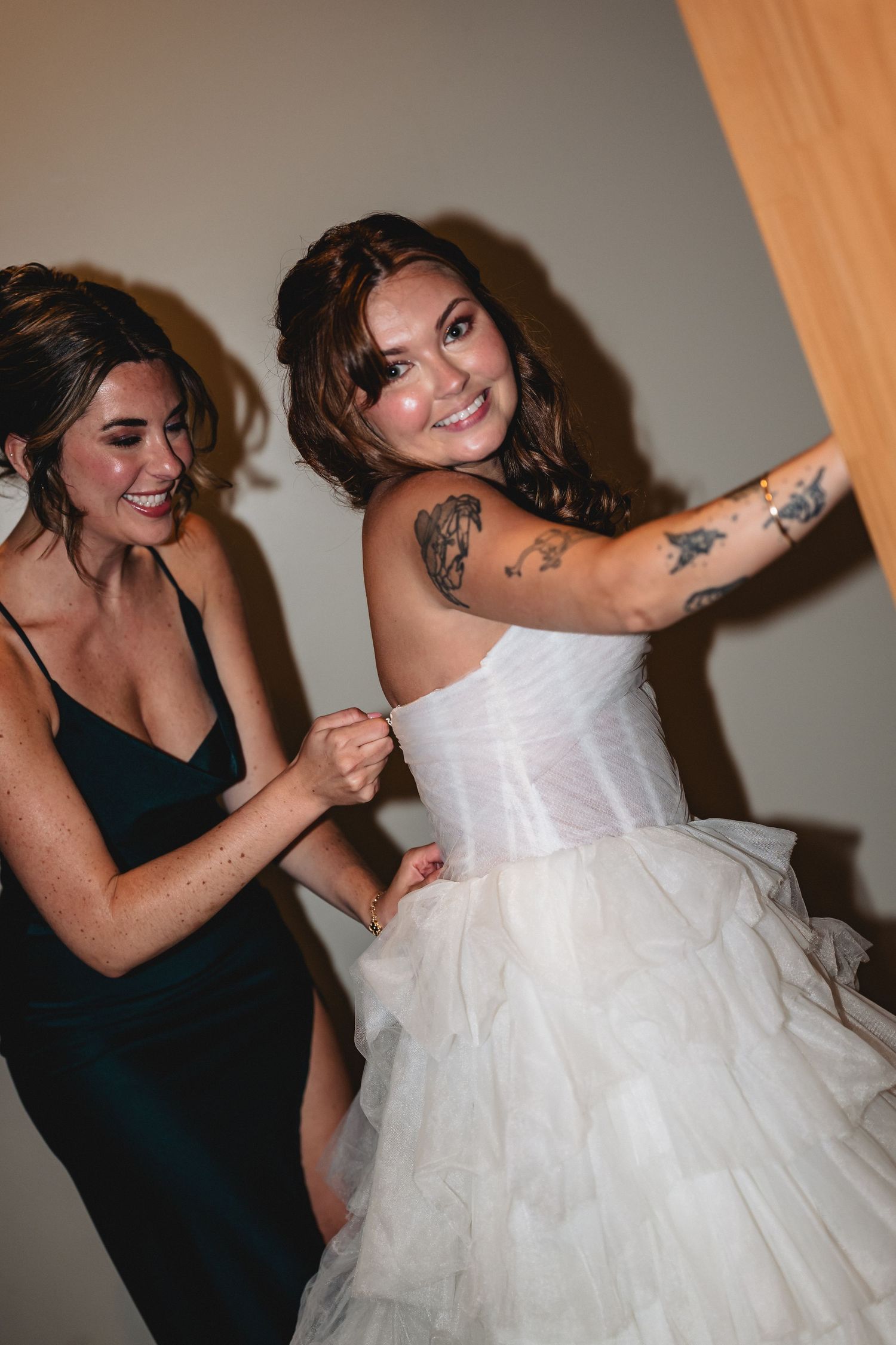 A bridesmaid in a dark green dress helps button up a tattooed bride in a white ruffled wedding gown.