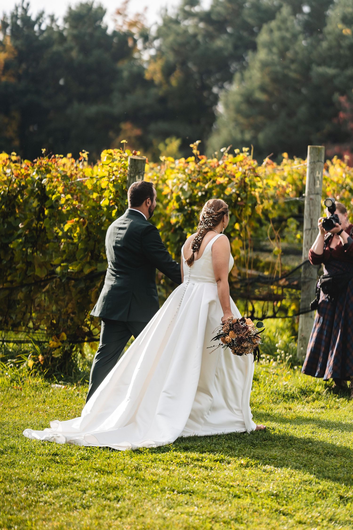 A bride in a white gown and groom in a dark suit walk through a vineyard during their fall outdoor wedding ceremony.