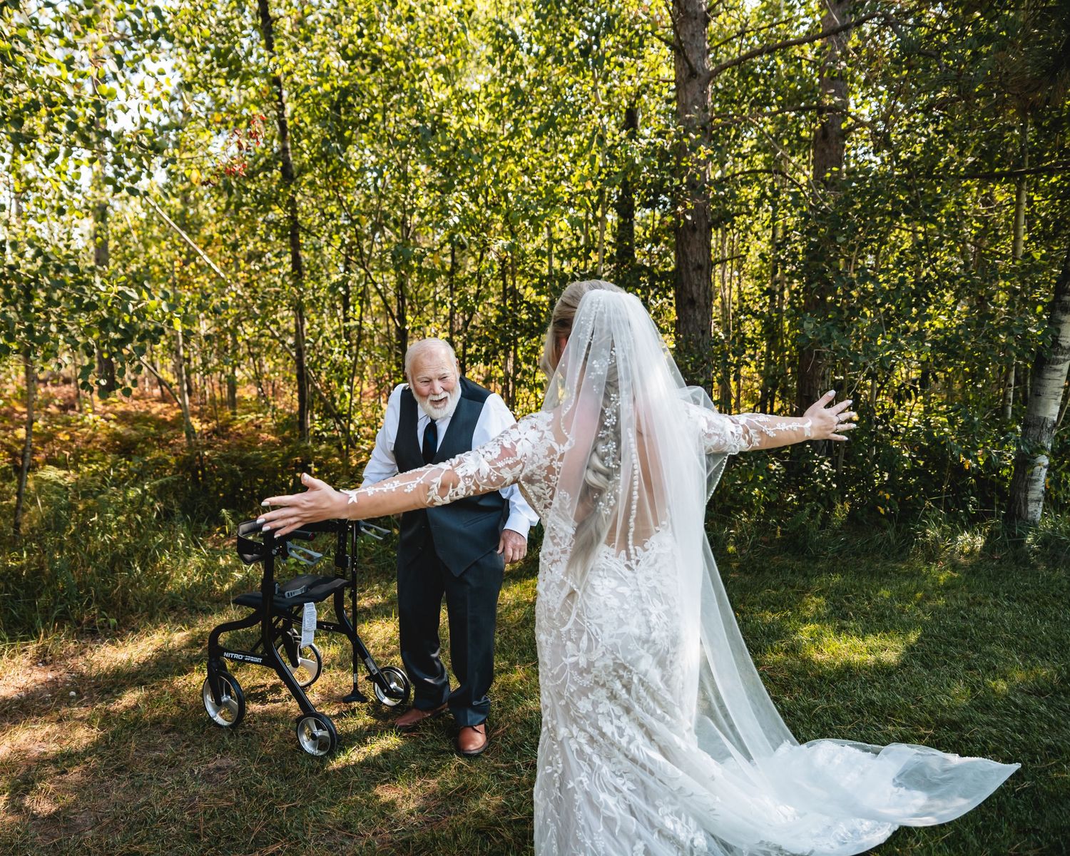 Elderly man with walker opens arms to bride in lace gown and veil in sunlit forest setting.