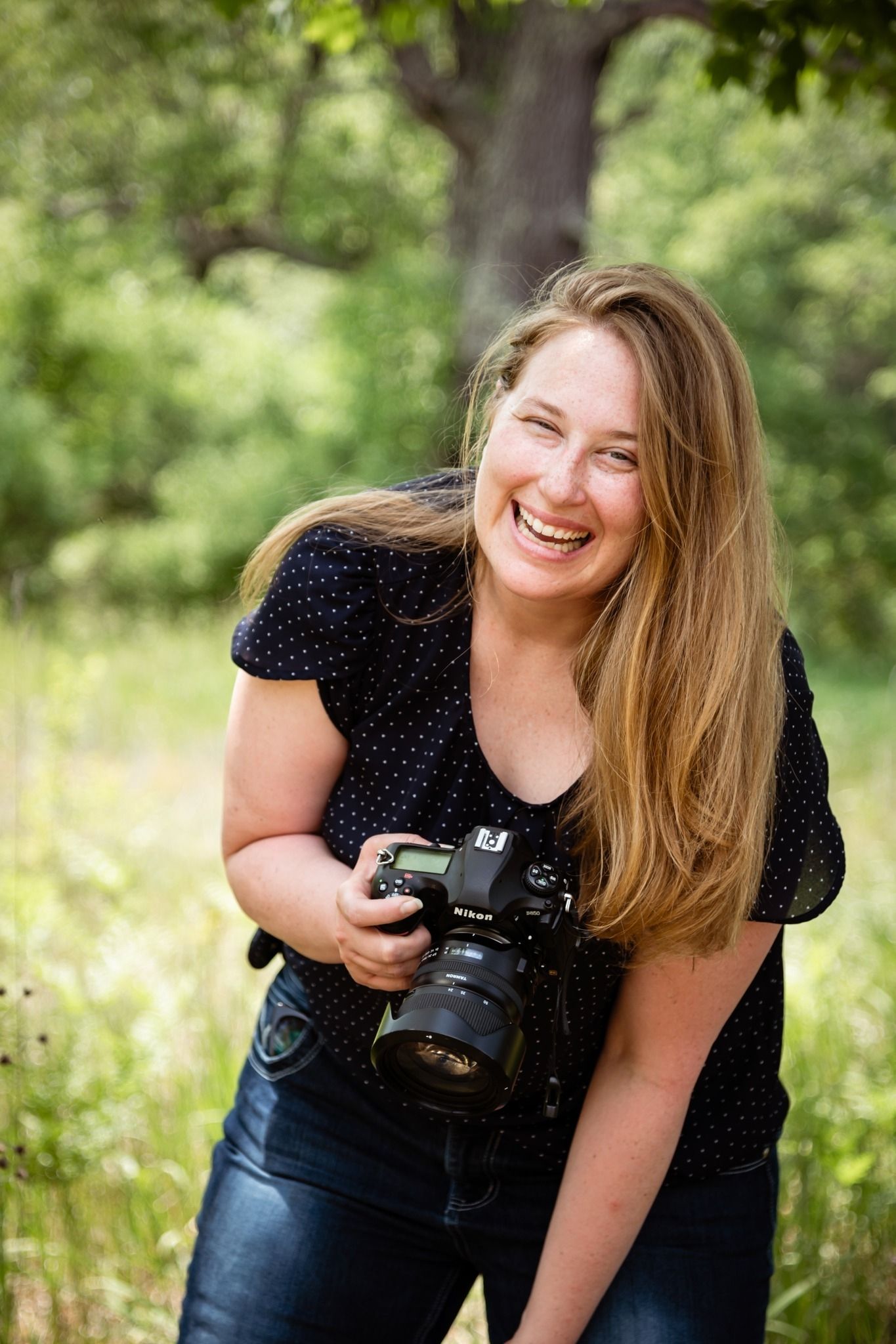 Photographer in polka dot top holds professional DSLR camera while laughing outdoors in nature setting.