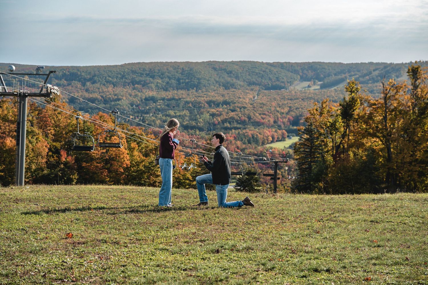 Man kneeling on one knee proposing to a woman on a grassy hilltop with a ski lift and colorful autumn at Boyne Highlands