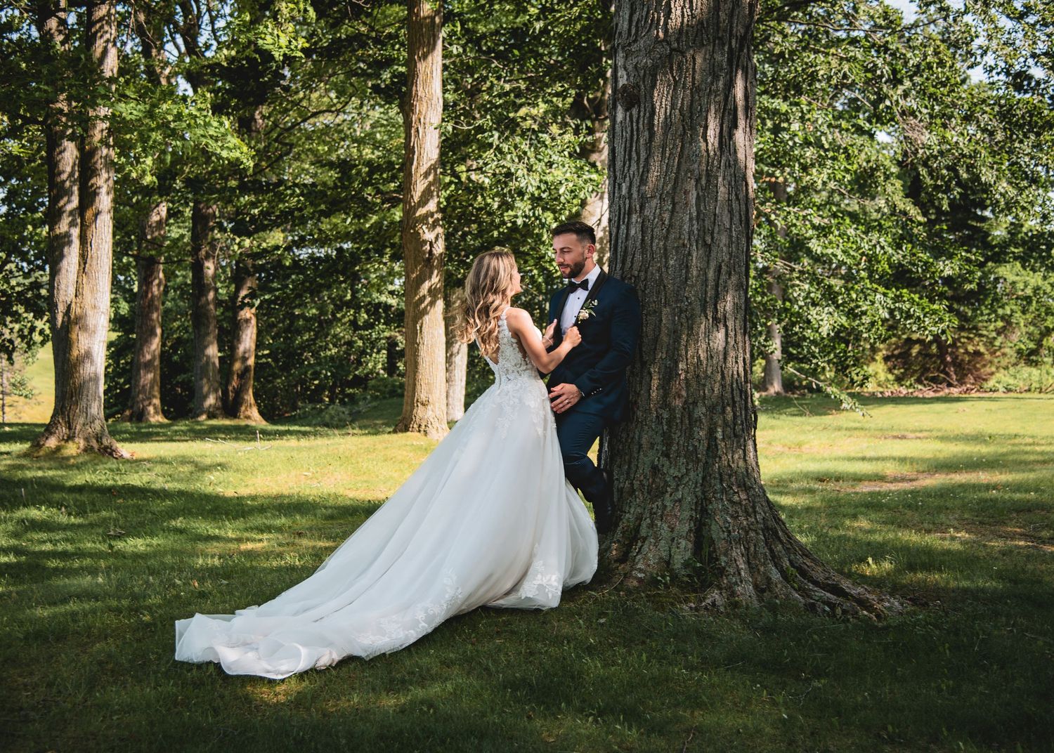 A couple in wedding attire pose together against a large tree trunk in a sunlit wooded area.